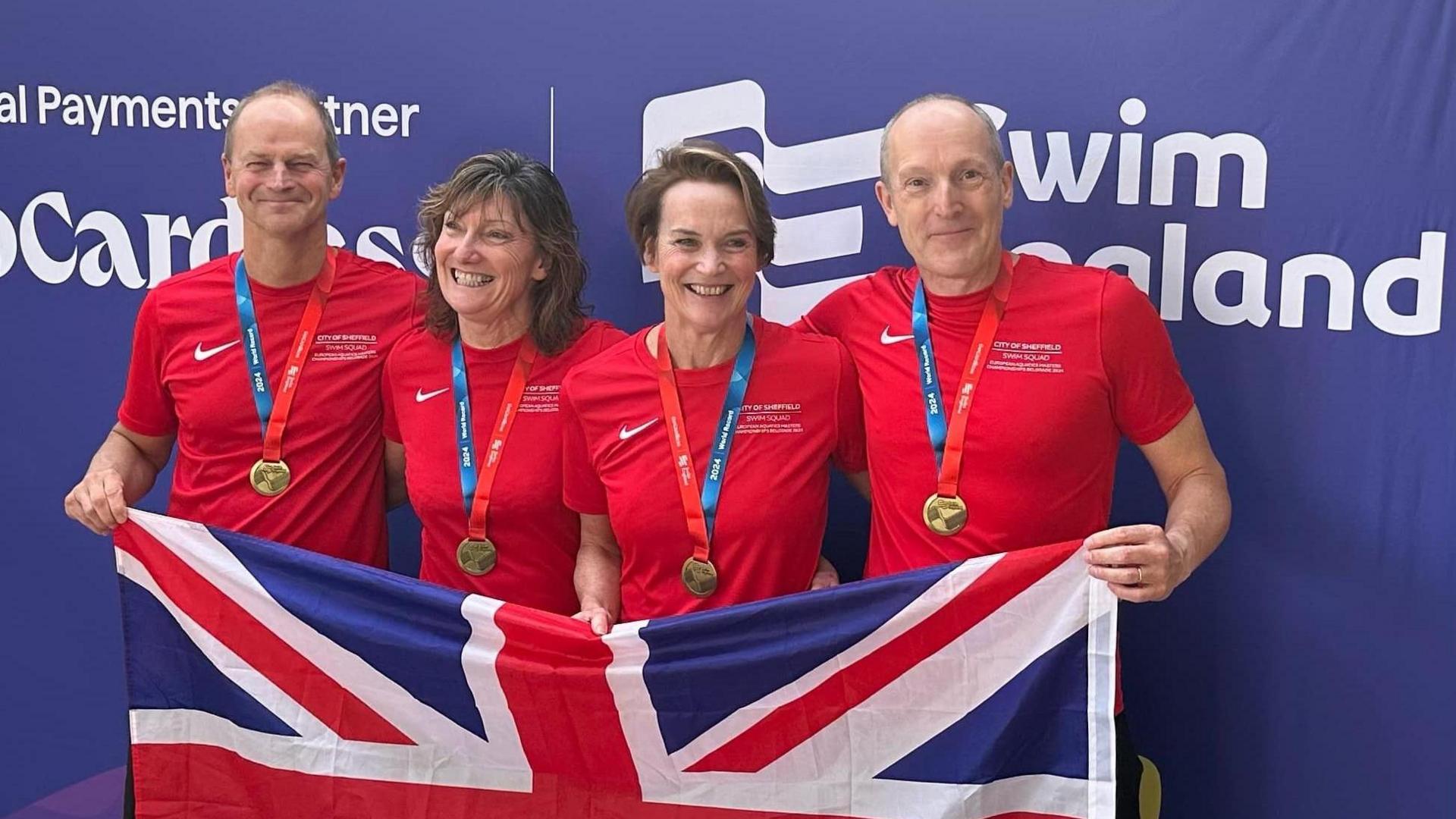 Swimmers Jonathon Saunders, Dawn Kissack, Lindsey Gaywood and Andrew Moore wearing red T-shirts and gold medals holding a Union Jack flag. 