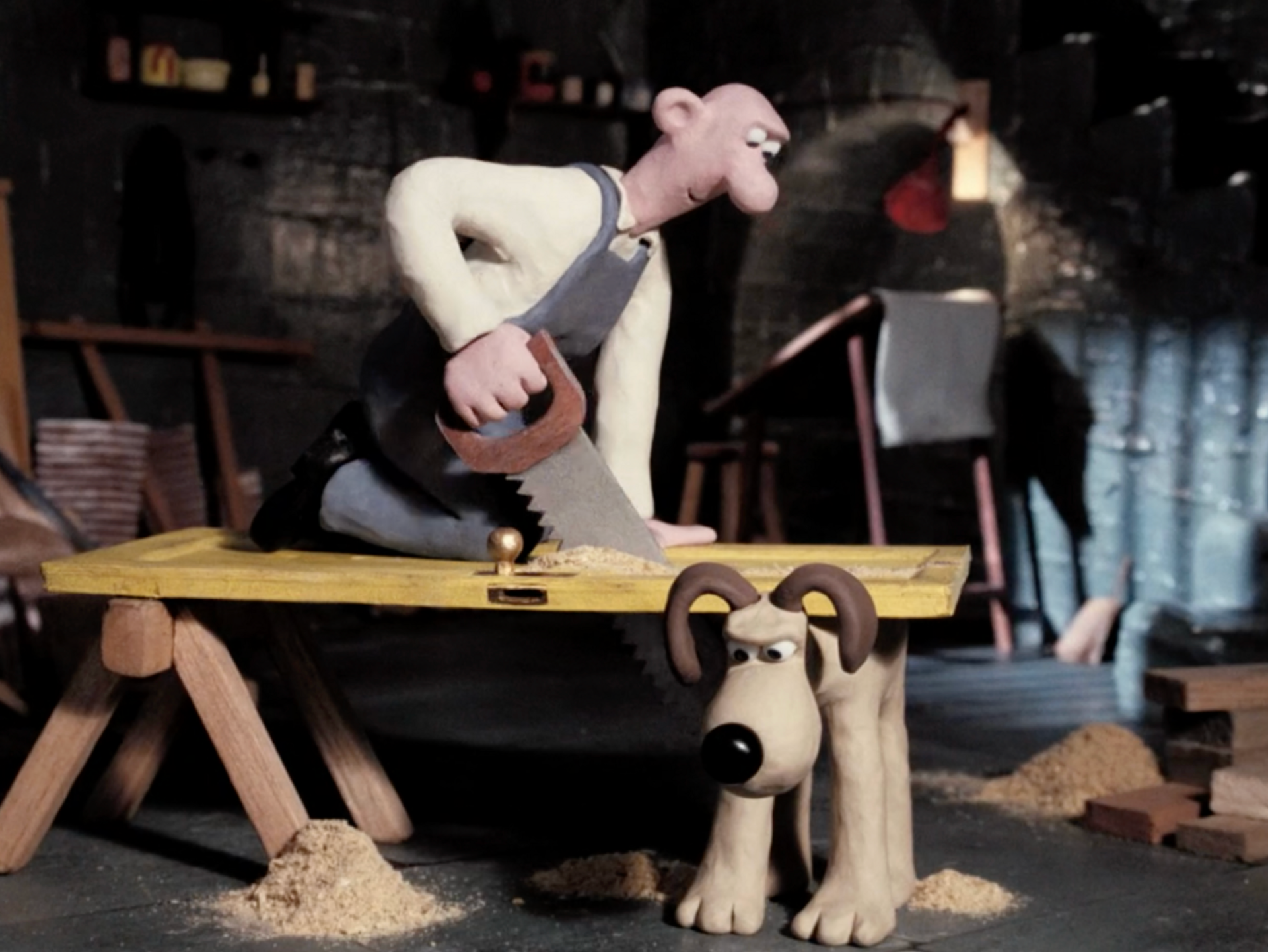 wallace cutting through a door with a saw on a trestle table. one set of the trestle table's legs is missing, so gromit is being used as a replacement