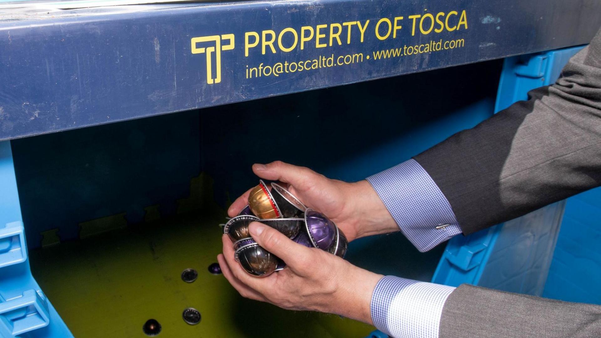 A man holding several coffee pods over a big, blue waste bin. He appears to be wearing a suit.