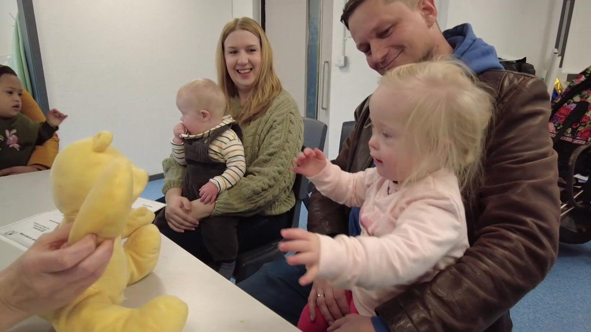 Three babies sat on their parents' knees at a table where they are engaging with a yellow teddy bear