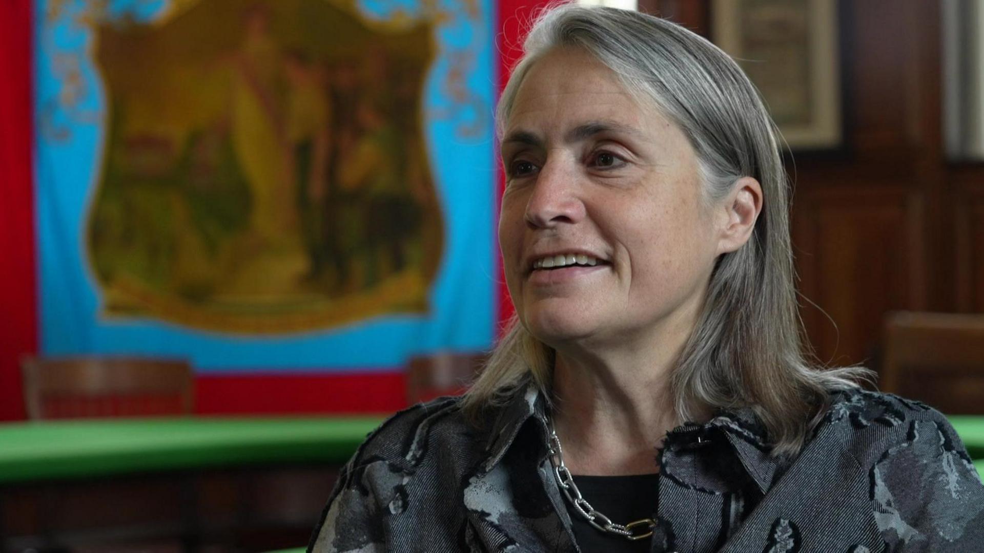 Fiona Hill smiling to the left of the camera. She has shoulder length grey hair and is wearing a grey shirt and silver chain necklace. She looks to be sitting in front of a blue and red miner's banner which a painting in the middle.