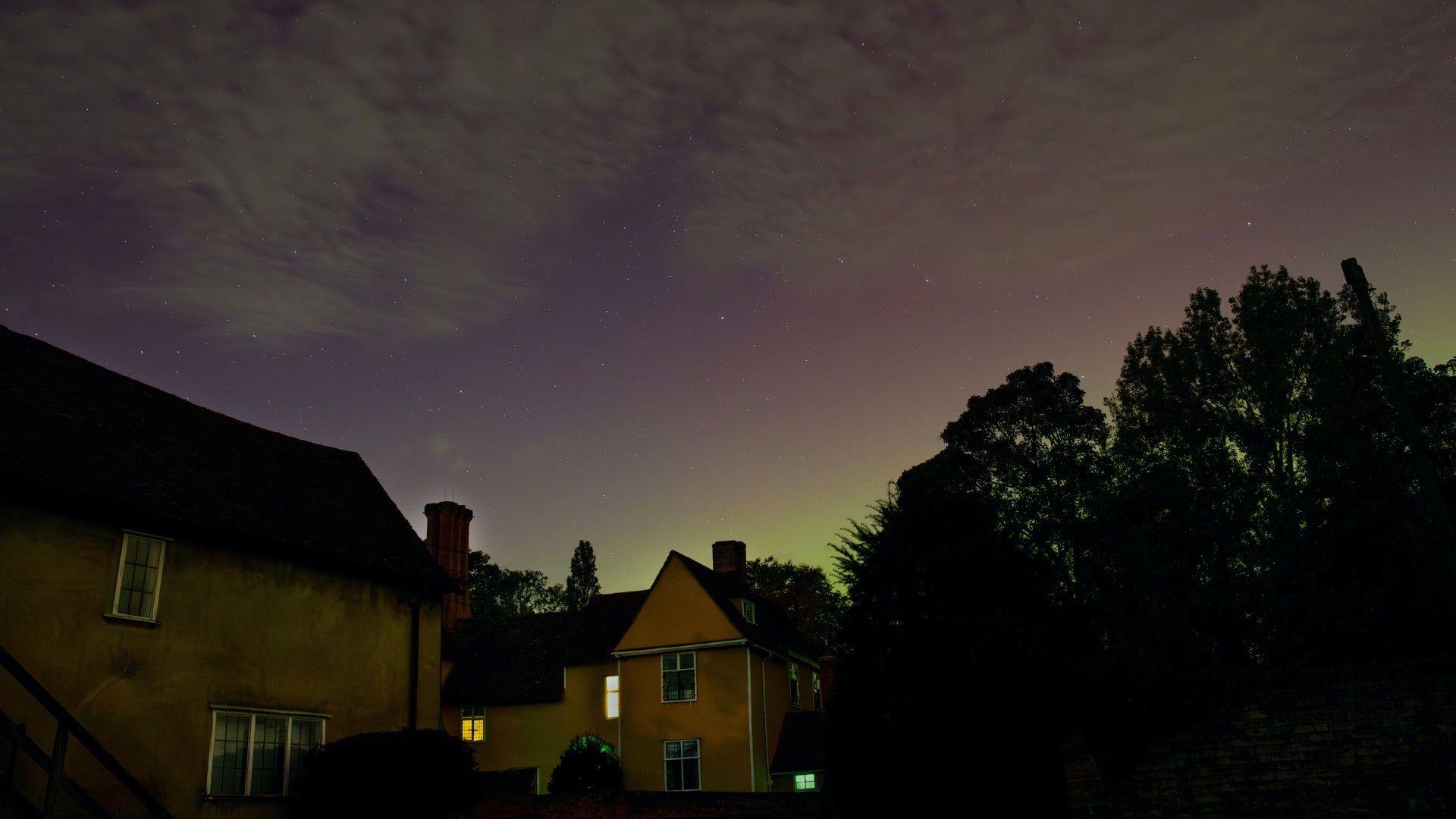 A picture taken at night of two buildings and some trees against a sky with faint green northern lights and scattered clouds.