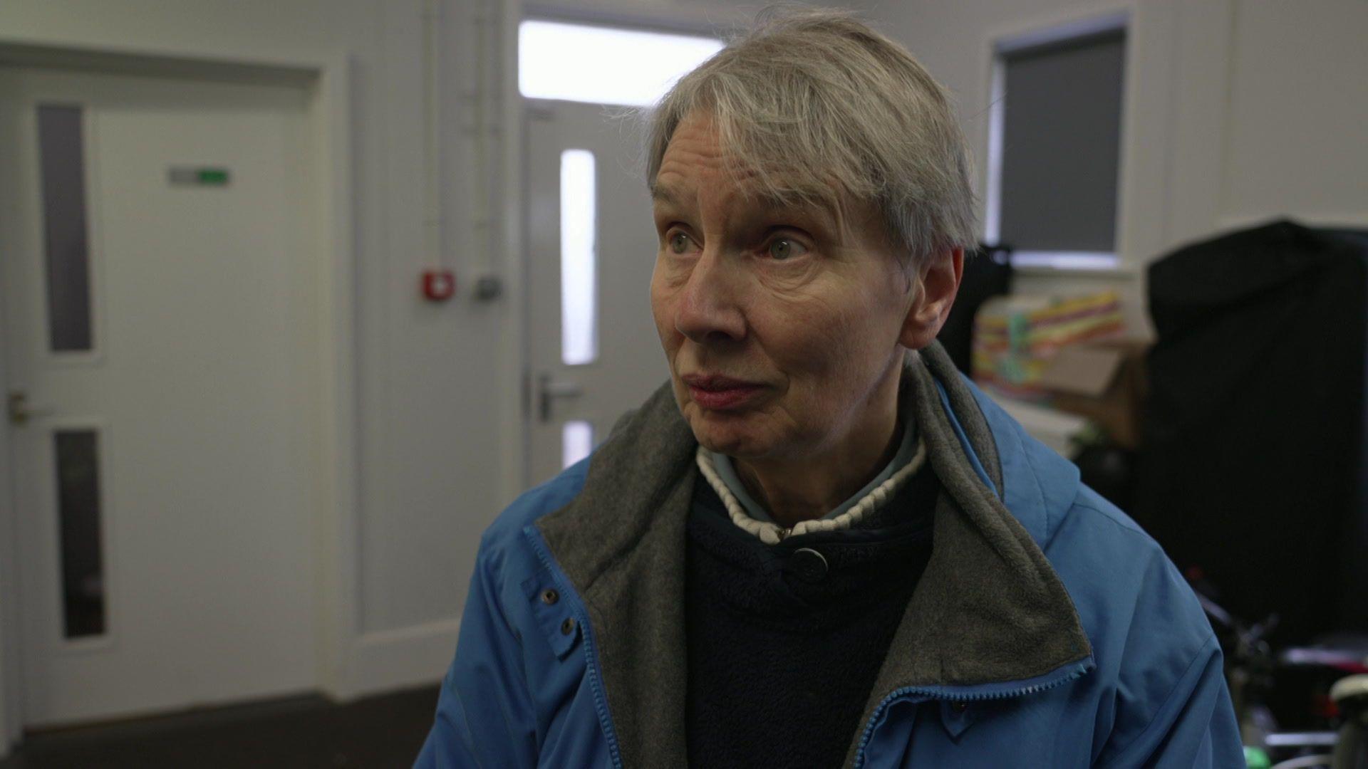 An elderly, white woman, looks to the left of the camera as she stands inside a community centre, wearing a blue coat and brown black fleece. She has short, grey hair