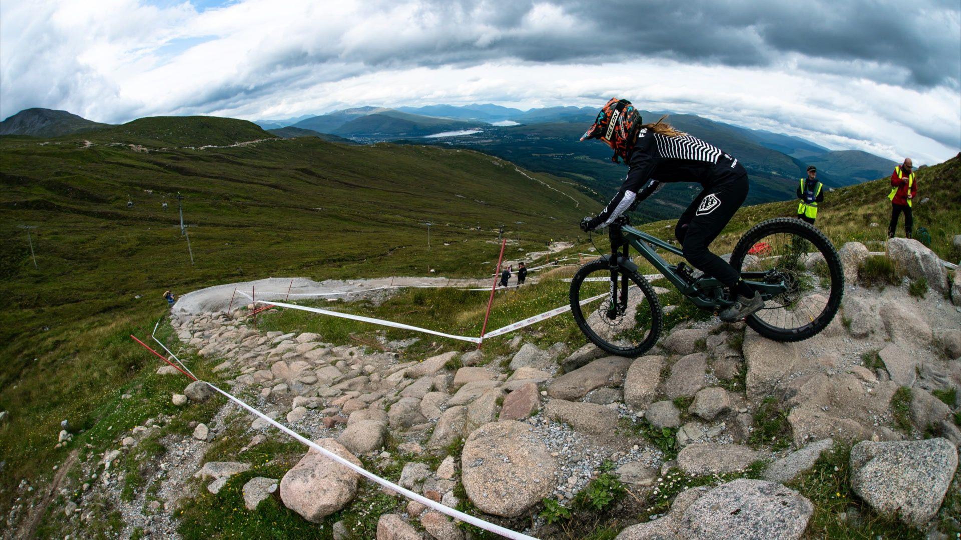 A mountain bike rider in a black outfit with a red helmet riding down a cobbled path with lots of green mountains in the background.