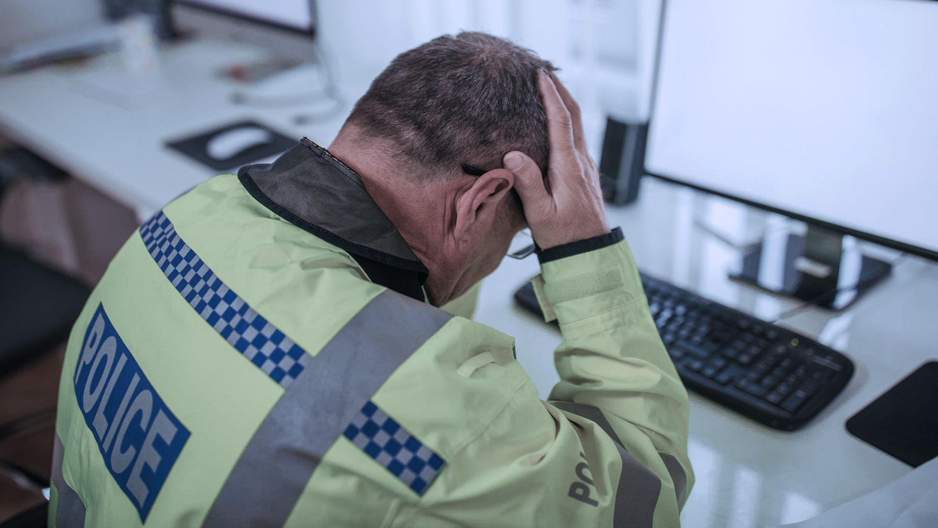 A police officer holds his head in his hands. He is an older man sitting at a desk. He is wearing a florescent yello jacket and glasses. The jacket has Police written on the back.