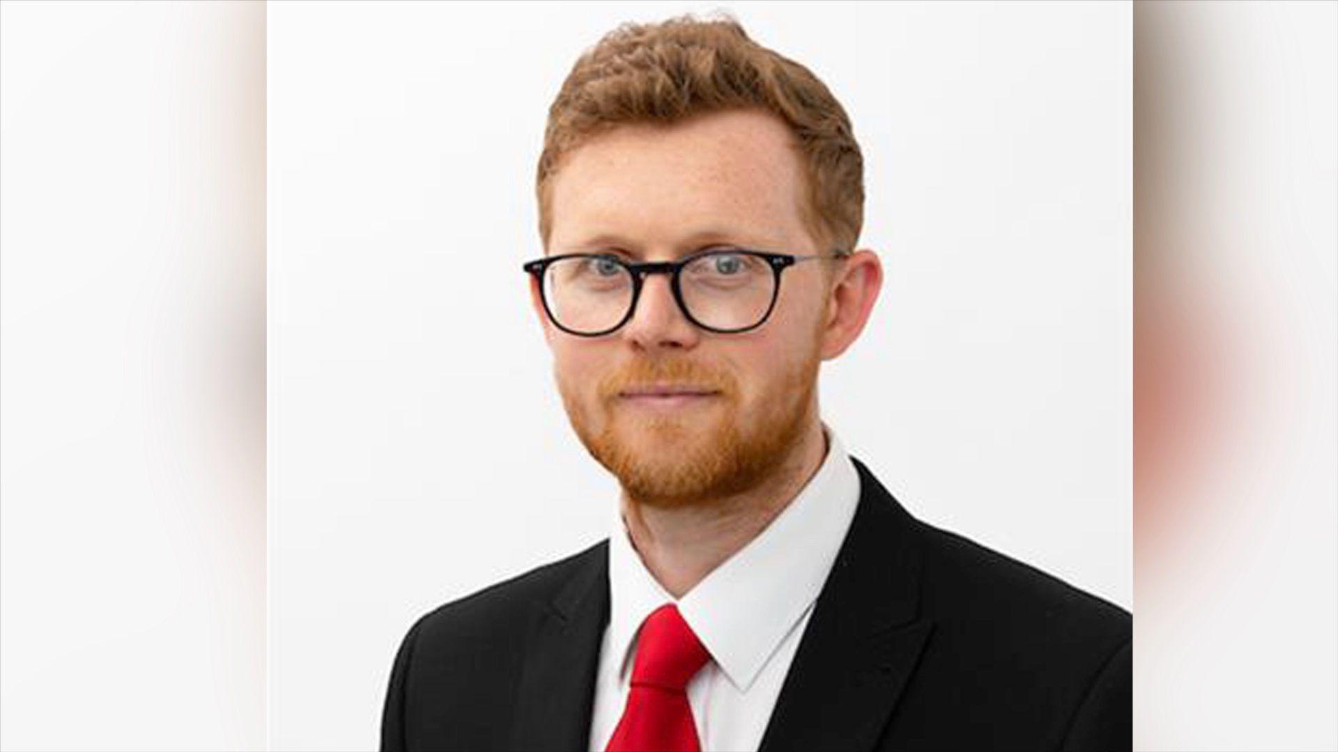 Headshot of Sam Gould in suit and red tie wearing glasses