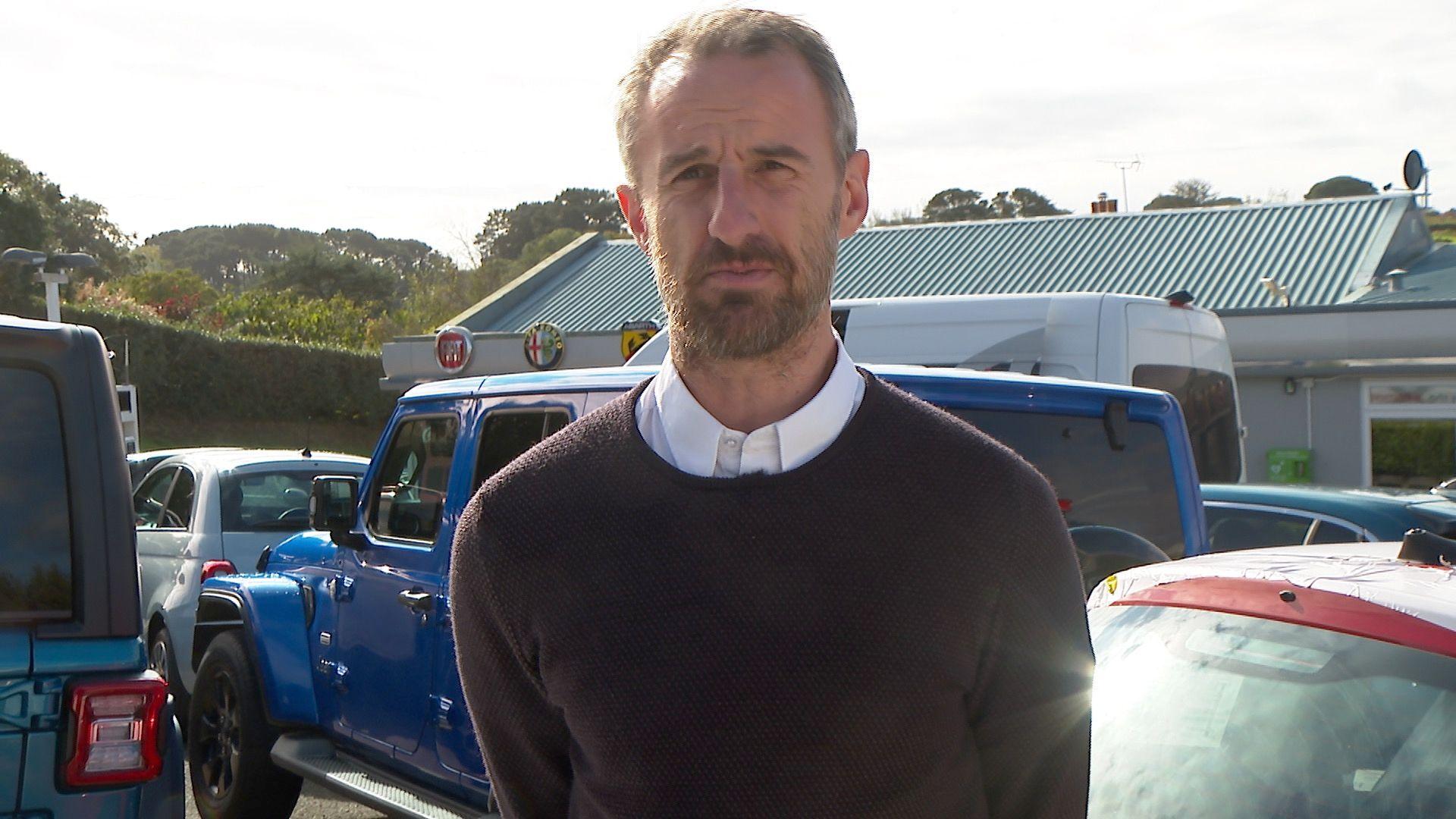 A man with a beard in a white shirt with a navy blue jumper on stands outside a car dealership.