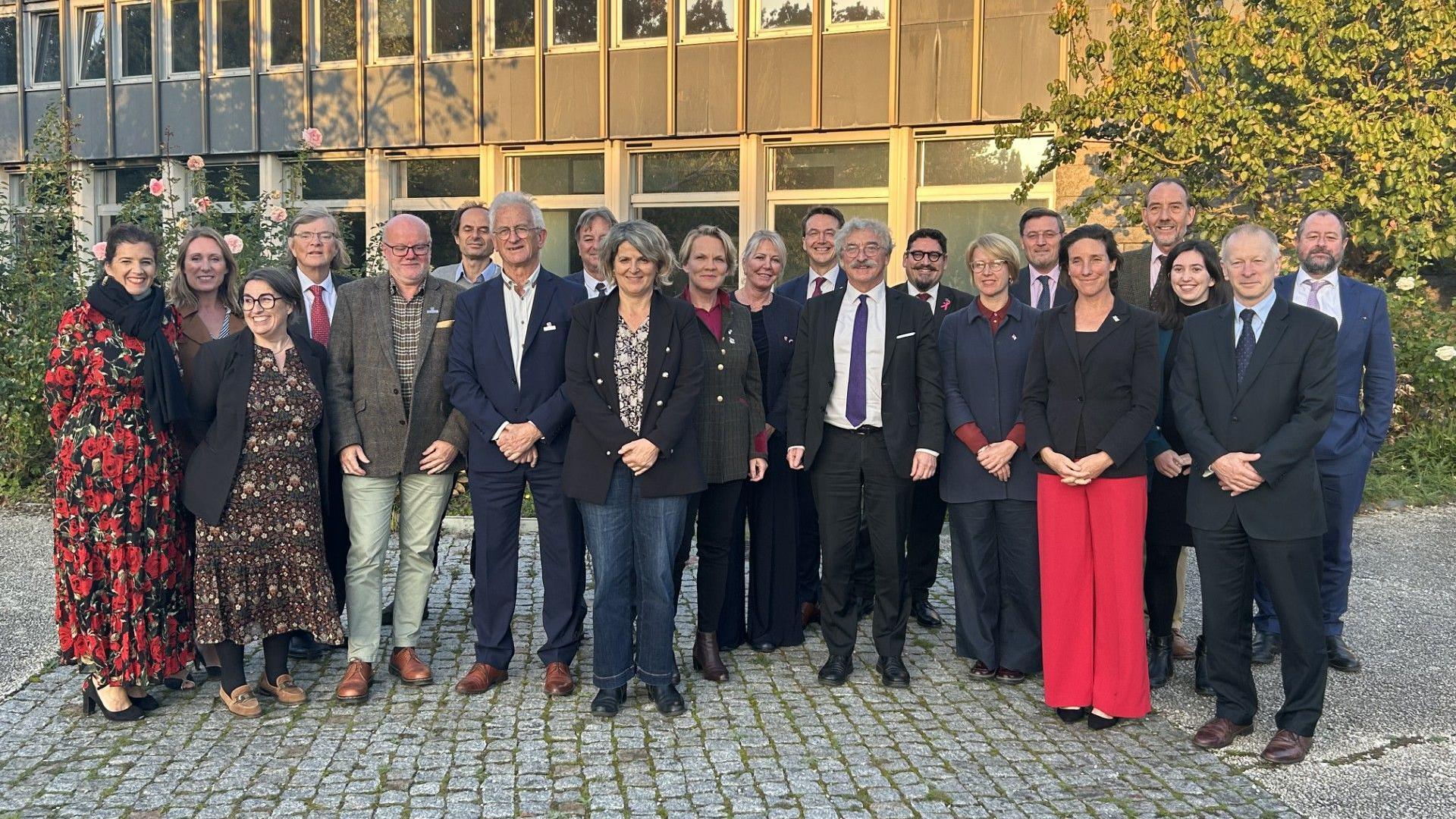 Men and women wearing suits and dresses and looking at the camera. They are all stood up. Below them is grey cobbled stone. Behind them is a grey building with glass windows. There are trees behind them on the right and a bush with pink flowers behind them on the left.