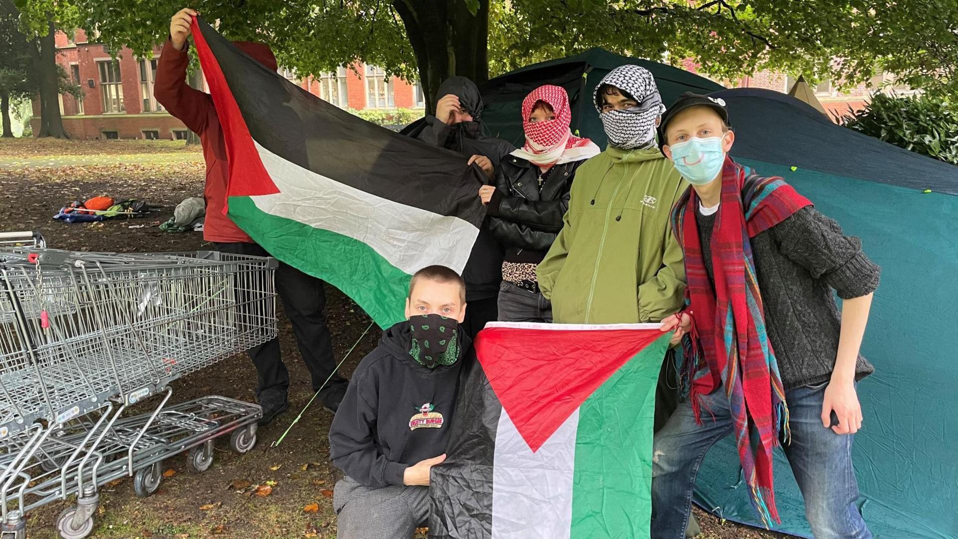 Six activists, all of whom are wearing face coverings or scarfs, hold two Palestinian flags next to a big, green tent. Two shopping trolleys are stationed next to the group.