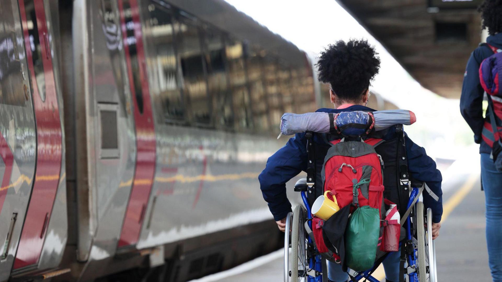 A woman in a wheelchair moves along a train platform with luggage in tow