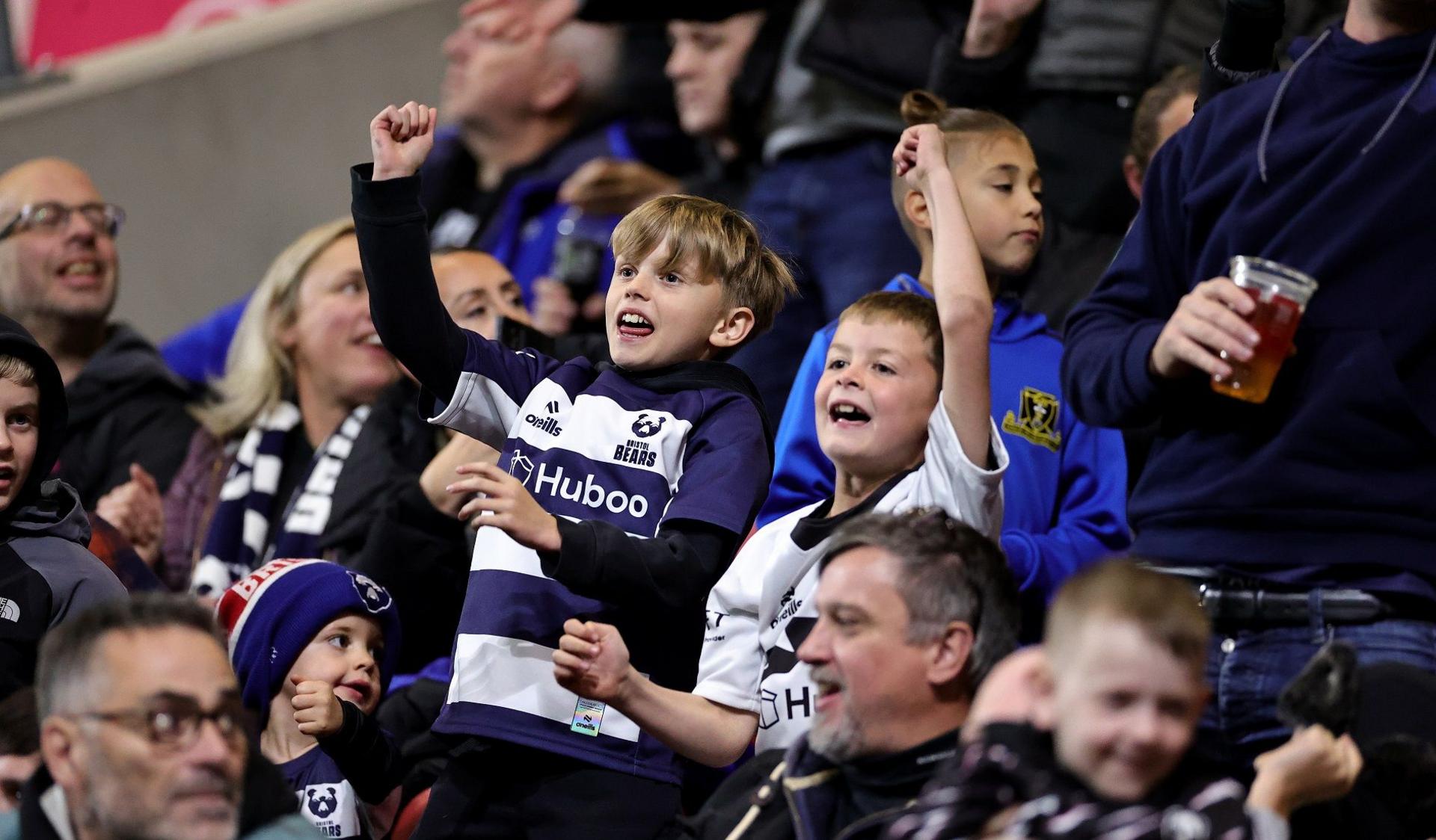 Two young boys in Bristol Bears kits cheer the team on during the match with Northampton at Ashton Gate. They are smiling as are many of the supporters around them