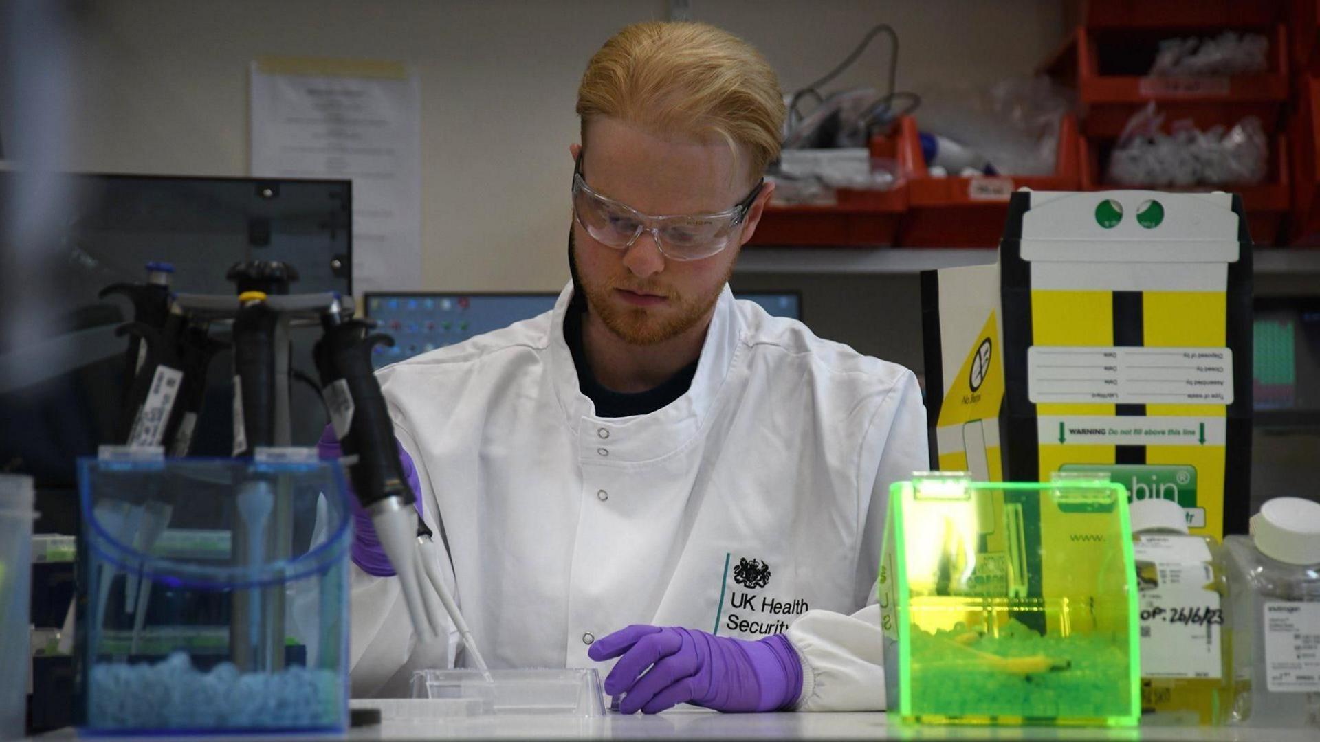 A lab technician wearing a white jacket, purple gloves and protective glasses while preparing samples. He has fair hair and is sitting at a desk.