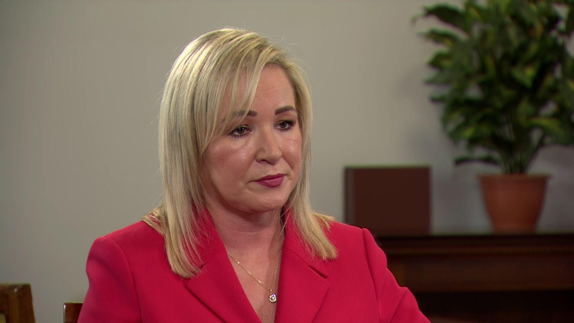 Michell O'Neill in a pink jacket with blonde hair. There is a plant behind her.