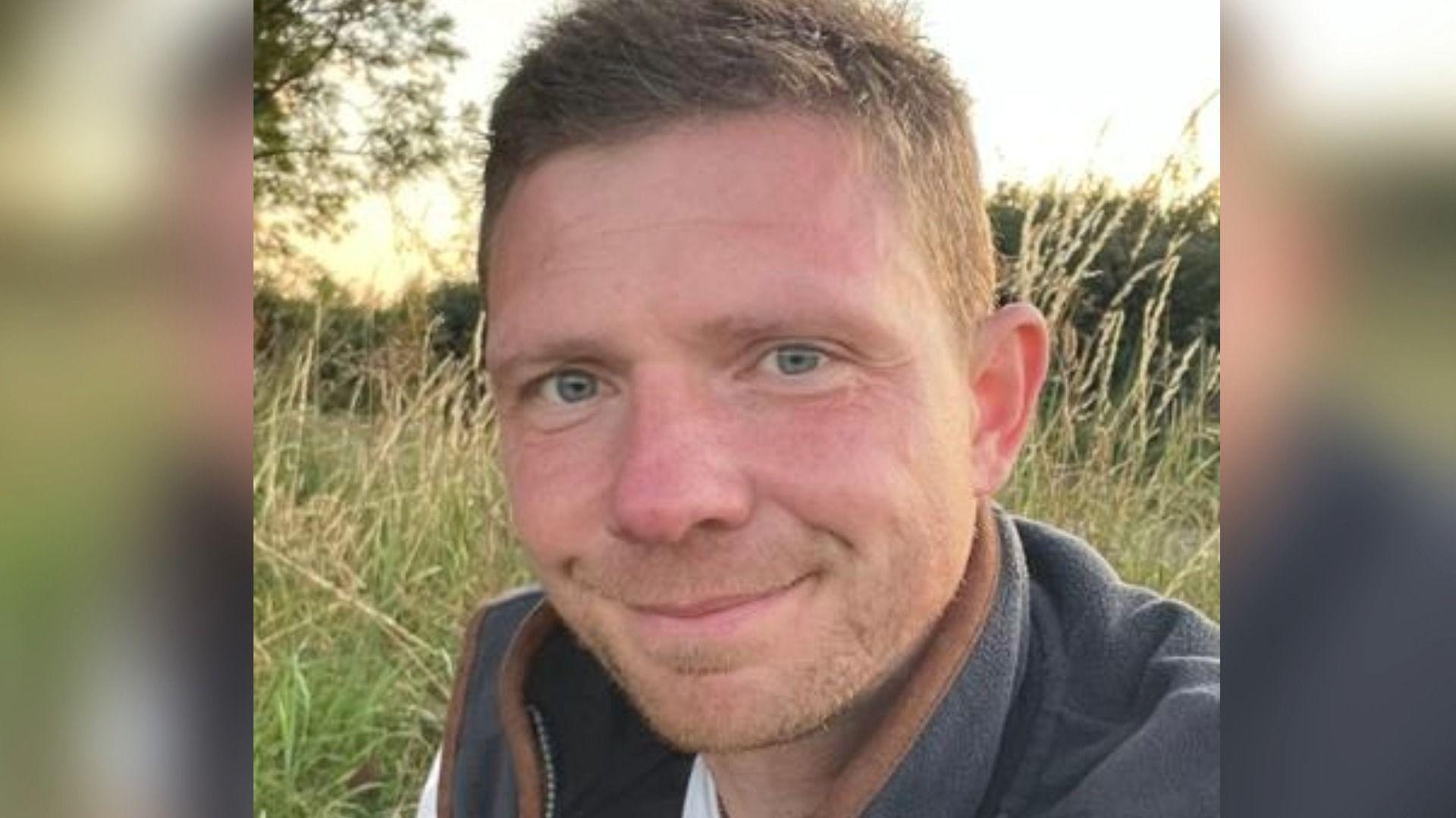 Dr Matt Clarke, who has short, dark hair and a light-coloured stubble looks at the camera, with a smile. He is wearing a grey fleece jacket, with a brown seam, and is standing in a field, with tall grass a hedge and a tree behind him.