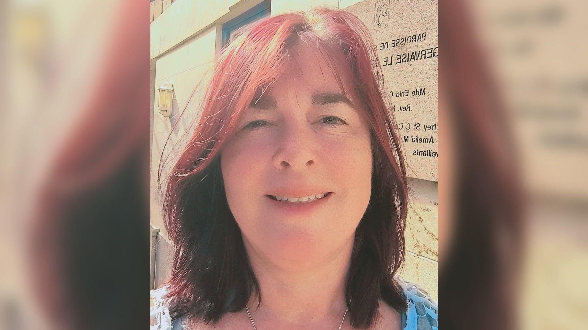 A woman with red shoulder length hair smiling, brown eyes wearing a light blue top, background of a granite building with an unreadable sign behind