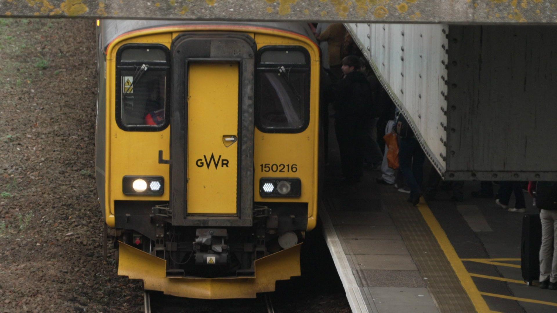 A train on the platform at a railway station. There are people getting on and off of the train from the platform. The train is yellow and black and says 'GWR' on the front.