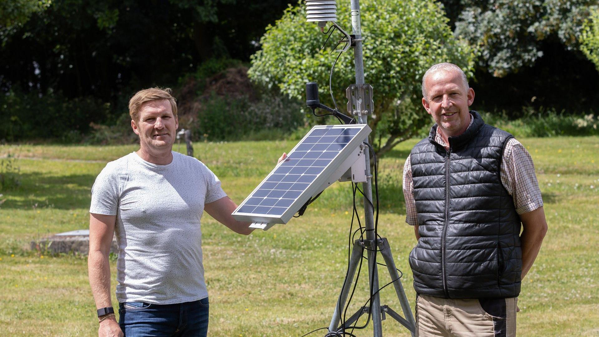 Two men stood in a green space with trees in the background by the weather station, which is taller than the men and formed from a tripod and pole with a solar panel and other attachments.