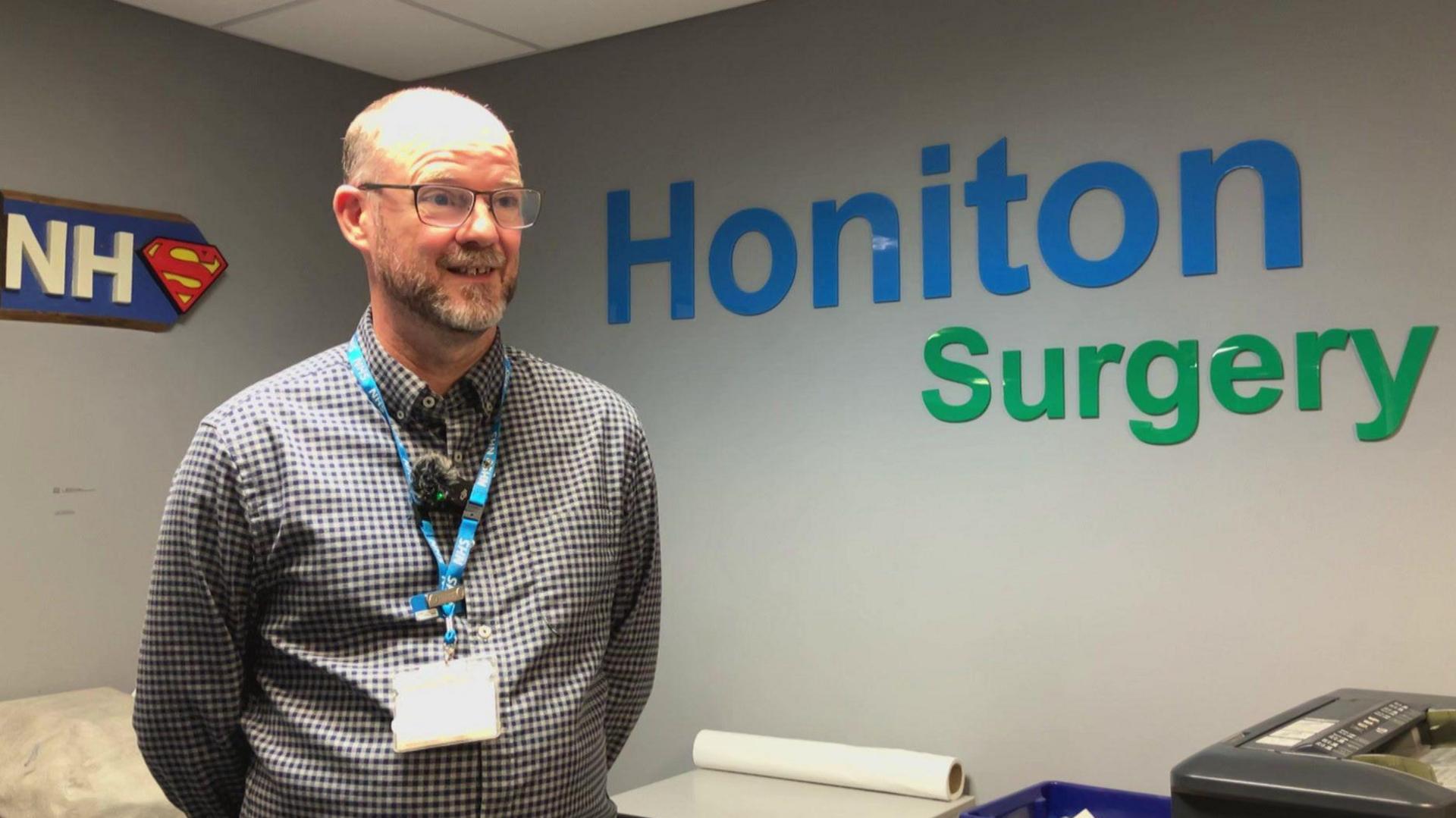 Chris Cruise stood to the left looking to the right. On the wall it has the words 'Honiton Surgery'. Chris is wearing a checked shirt and has a lanyard on.