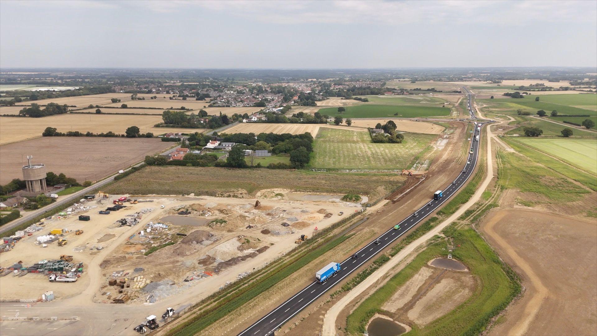 An aerial image taken by a drone looks down on a road with traffic using it, flanked by a roadworks site on one side and fields on the other, with a town seen in the background.