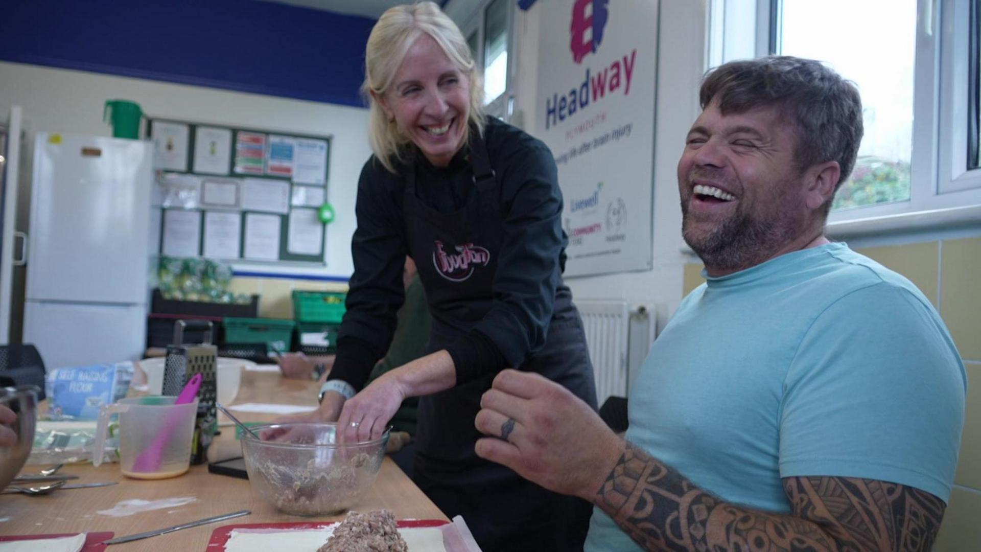 A man wearing a blue T-shirt laughing and smiling with a lady with blonde hair. The man has tattoos up his arm. The woman is wearing a black apron.