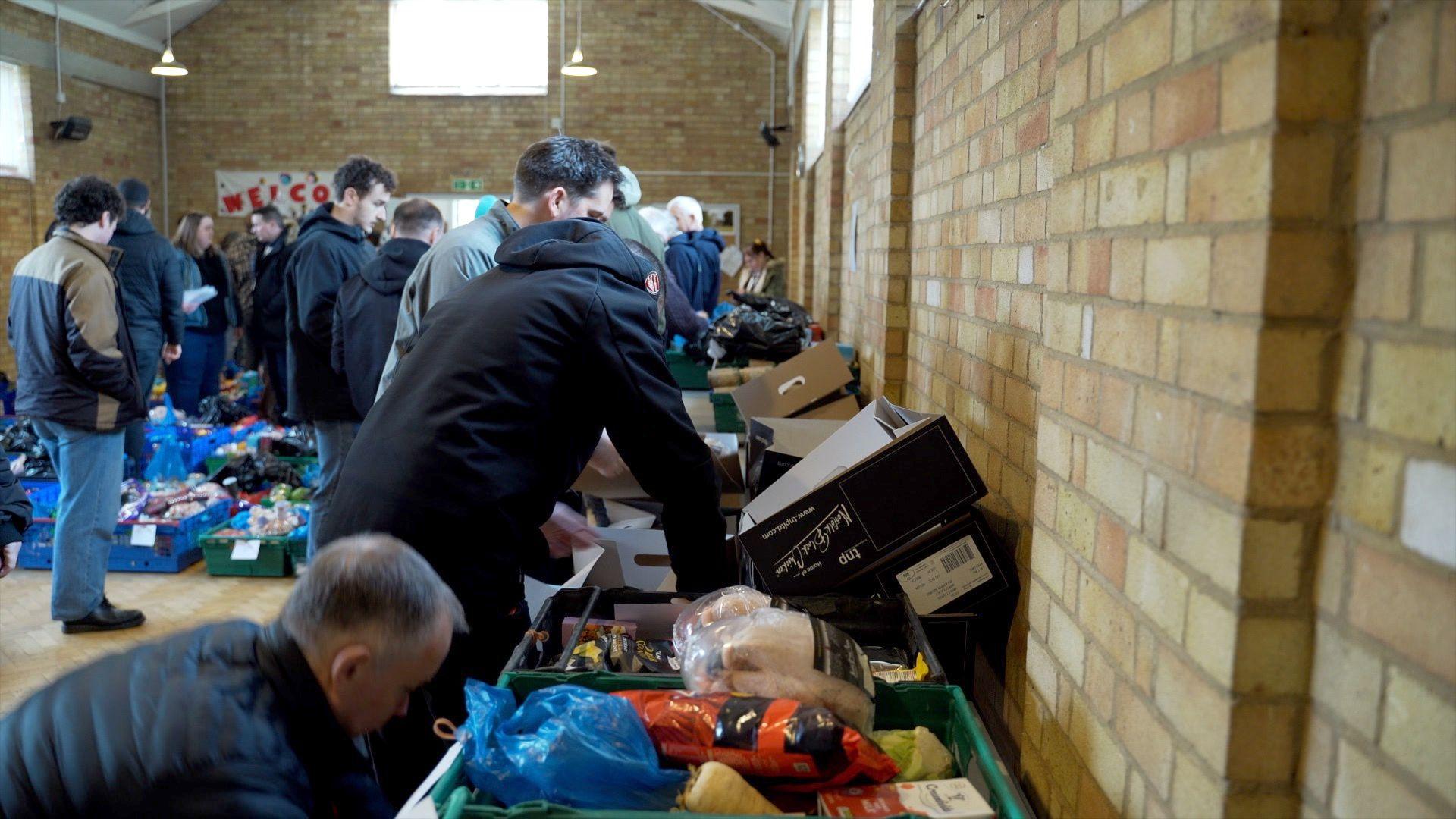 A large group of people in a community hall sort through food hampers