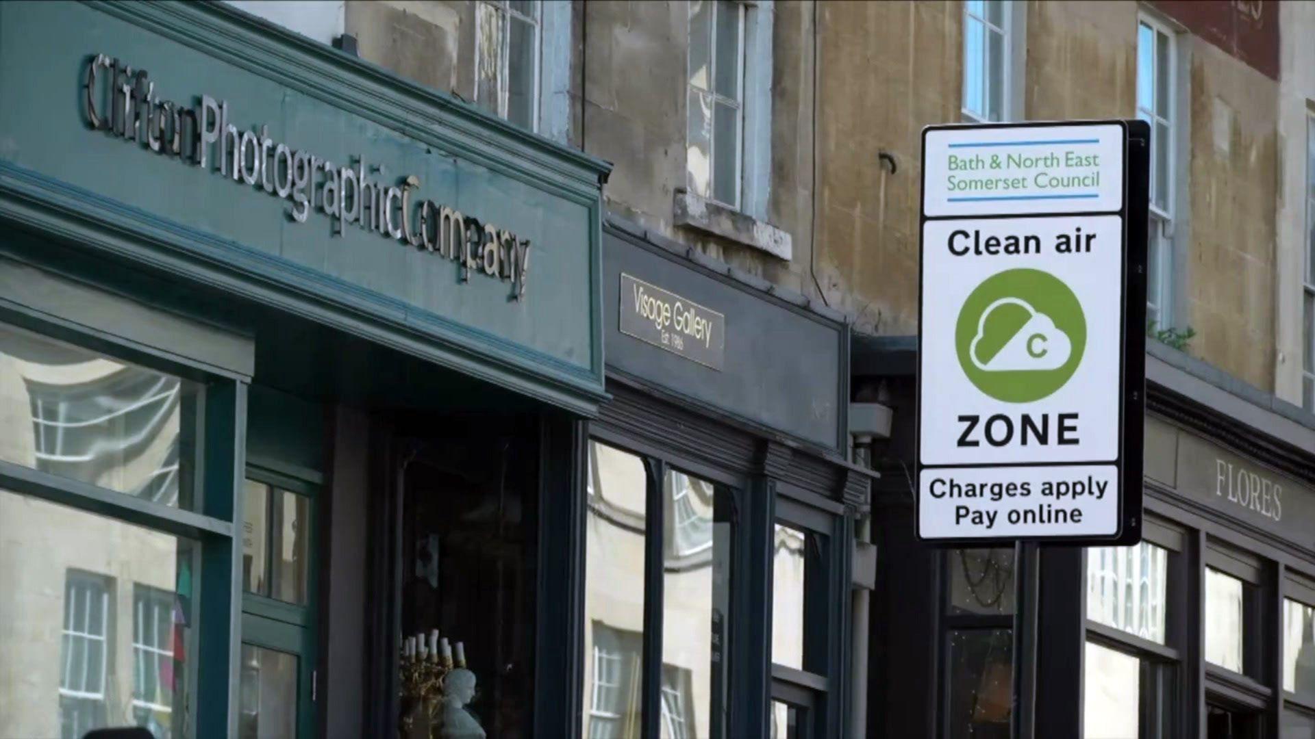 A sign marking the entry to the city of Bath's clean air zone beside shops in the high street. The sign is white with a green circle and a cloud on it.