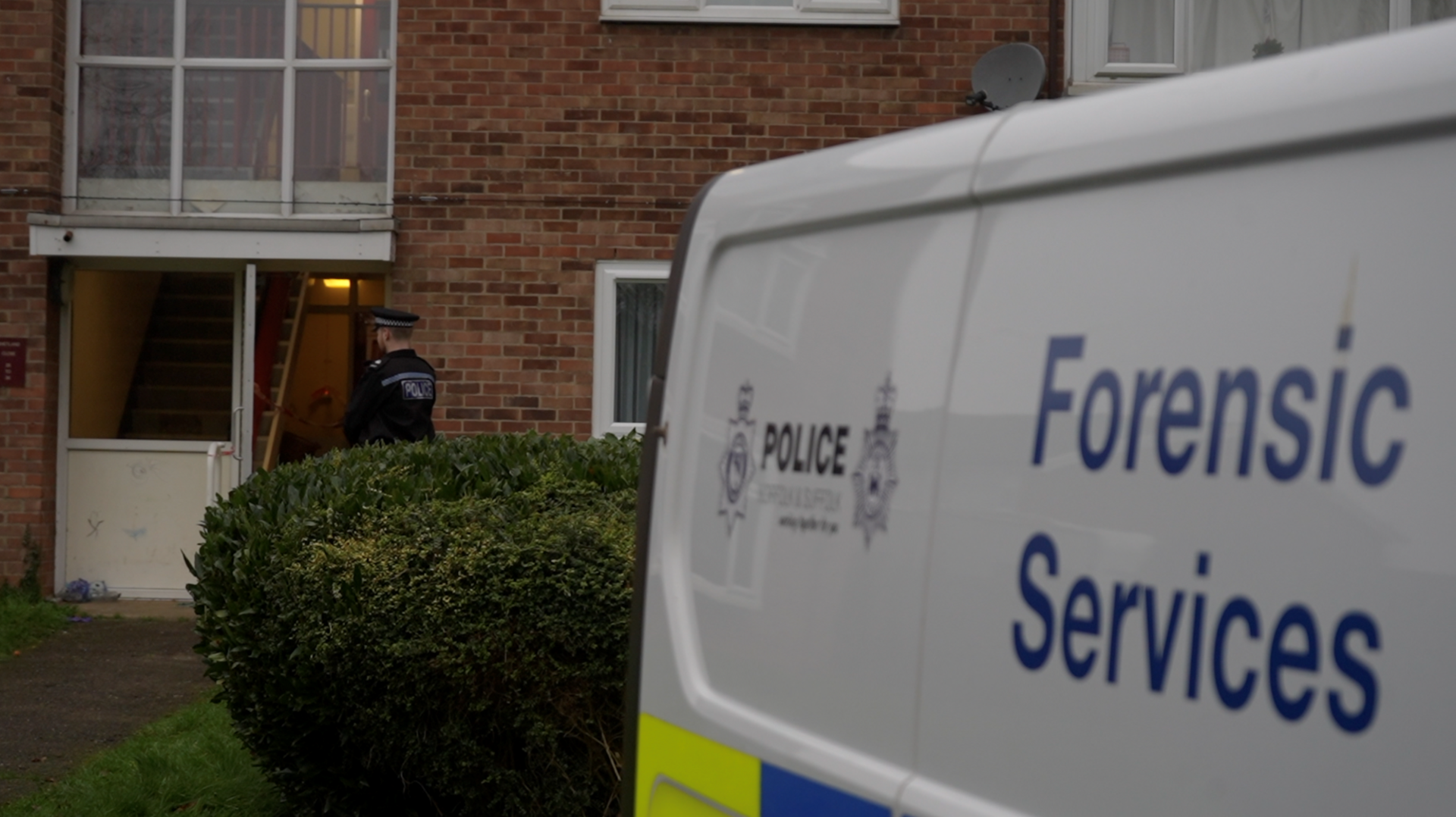 A white police forensic services van parked next to a building of flats. A policeman is standing next to the door. There is a green hedge between the van and the brick-built property. 