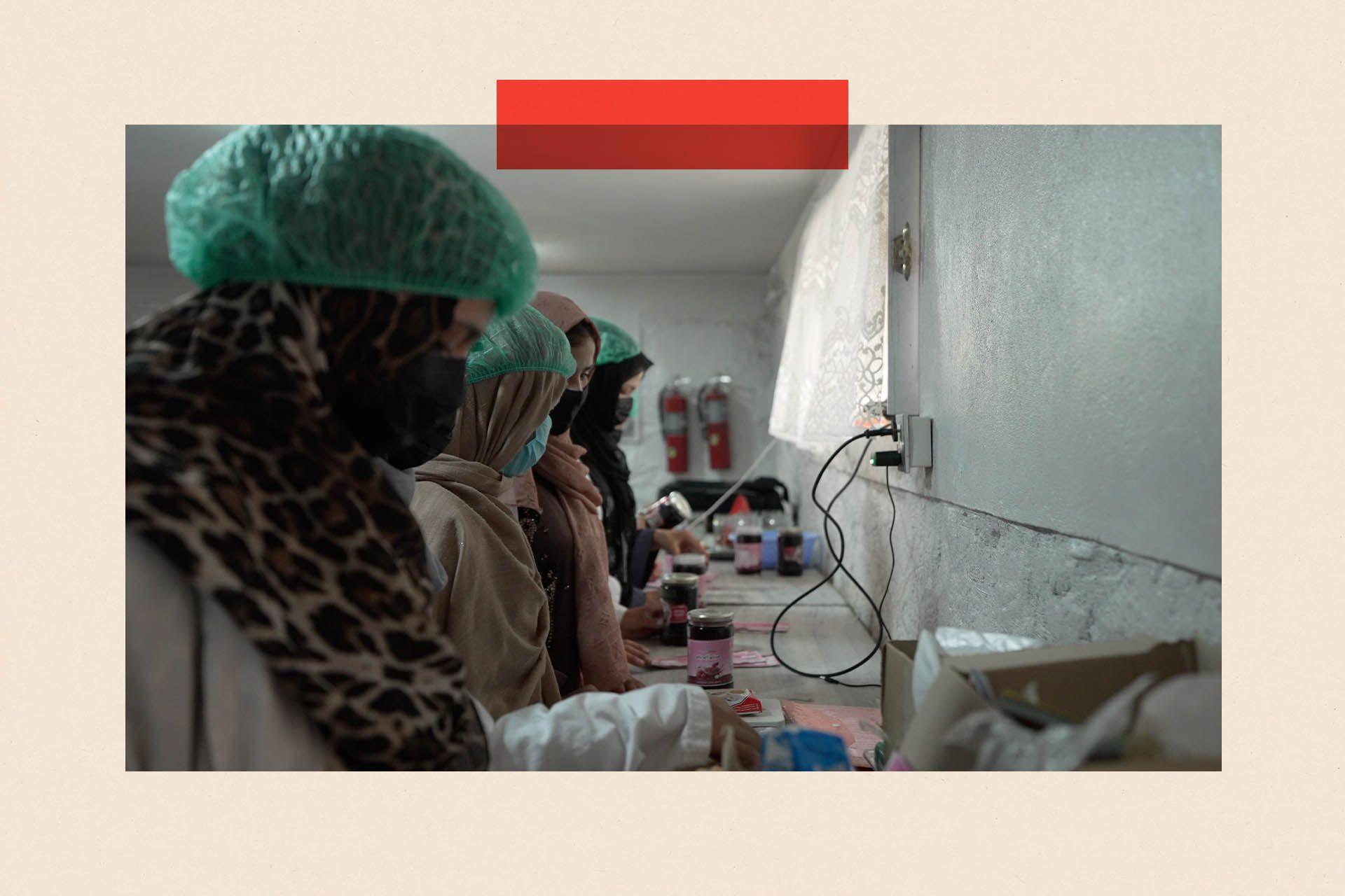 A row of women in headscarves and hair nets work at a long table with jam jars on it