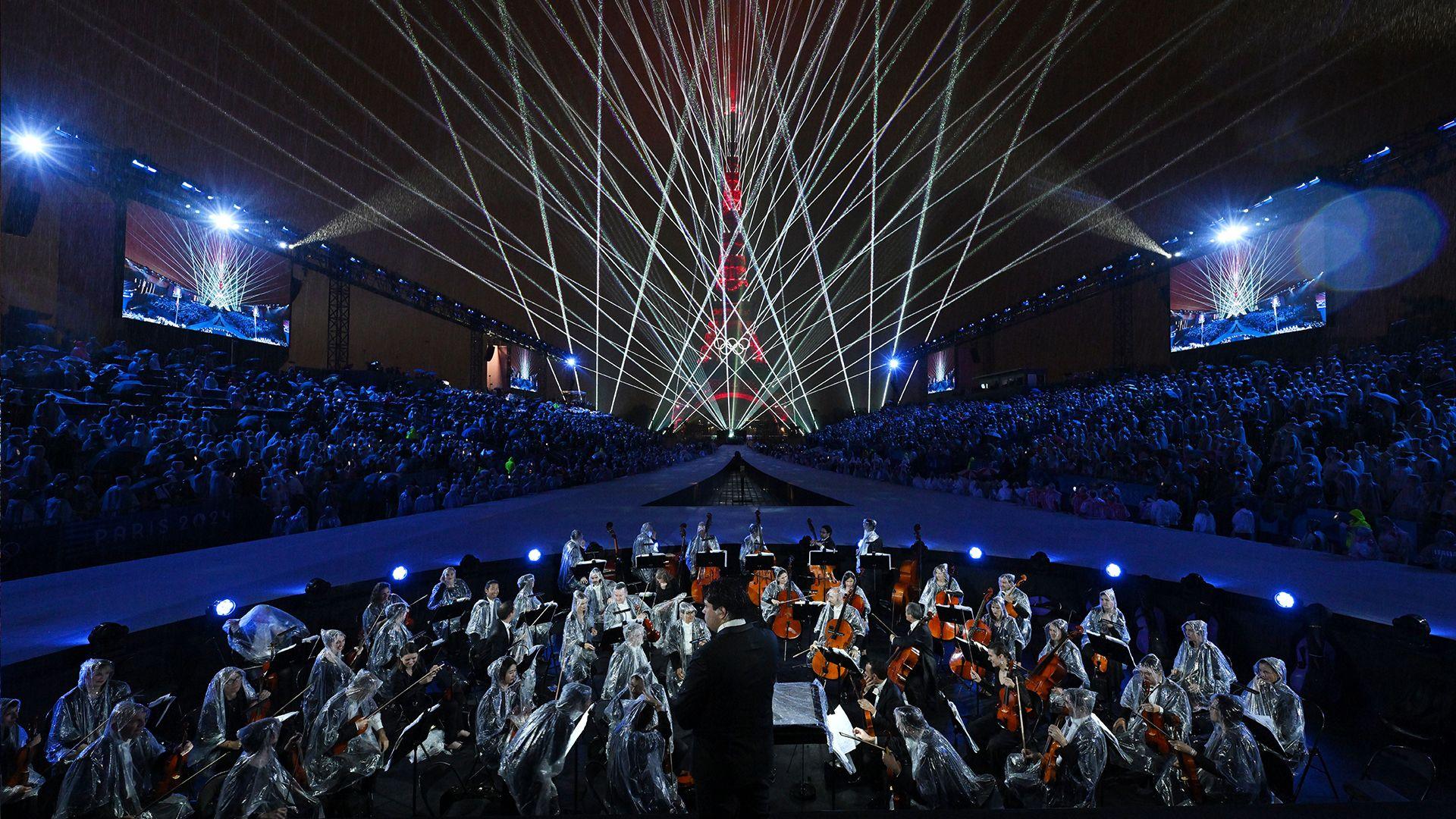 An orchestra wearing ponchos gets ready to perform in the rain, with a laser show behind them