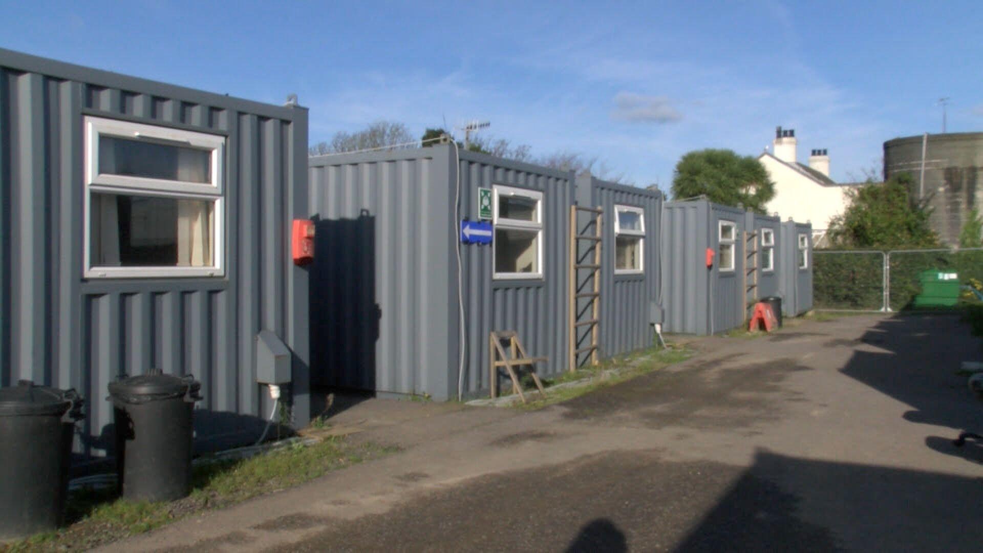 Three corrugated iron crates with windows in front of a row of houses.