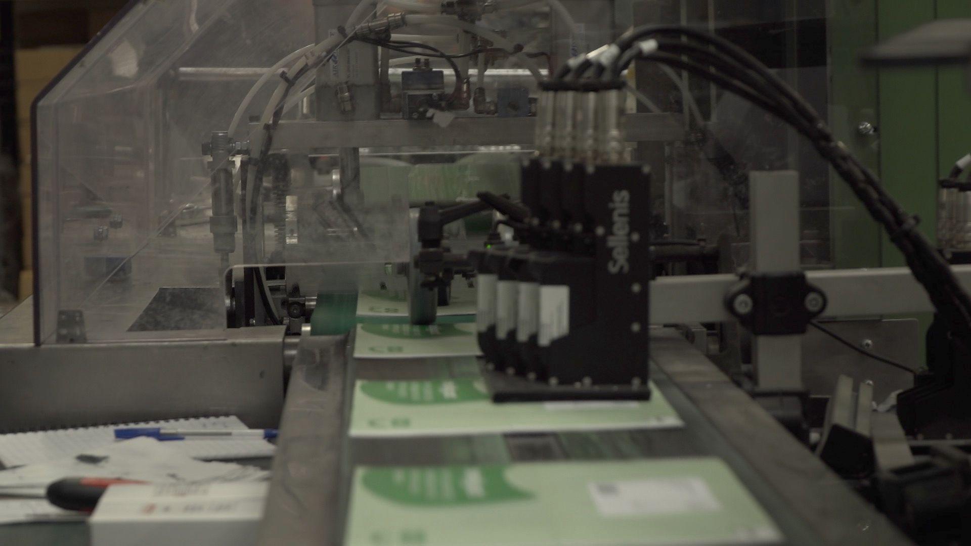 Green leaflets running through a machine inside First Mailing's premises. The production line is running towards the camera and the leaflets are mostly out of focus. A large transparent plastic canopy is further back, covering both the production line and some machinery inside. There are wires and tubes coming out of parts of the machines.