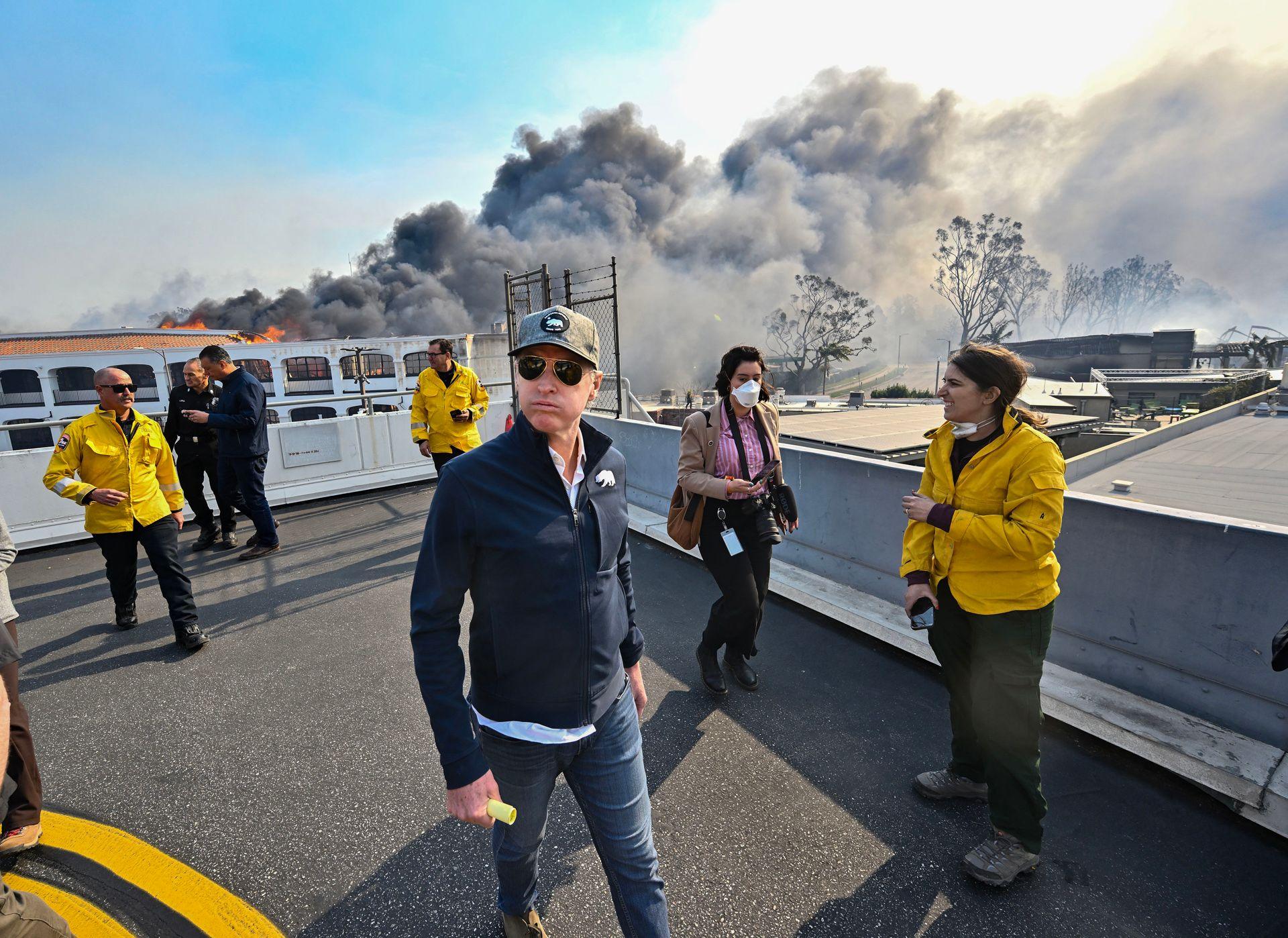 California Governor Gavin Newsom looks away from camera as he surveys damage during the Palisades Fire, surrounded by personnel and with smoke rising in the background.