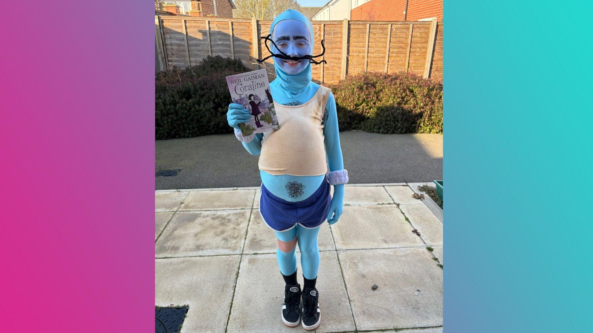 A girl smiling as she wears a blue morph suit with a beige vest and navy shorts over the top. Her hair is hidden by the morph suit and she's wearing blue face paint and a black moustache that's been made out of pipe cleaners. She has padding in the costume to make her tummy stick out and she's holding a copy of the book Coraline.