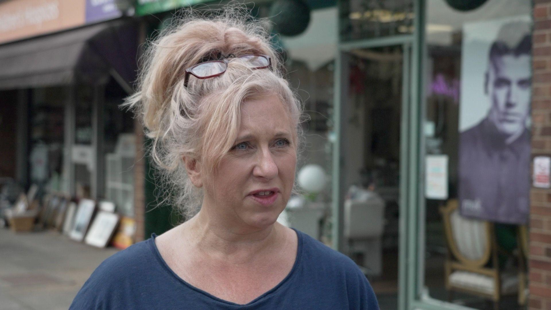 A blonde woman with glasses on her head is wearing a blue top as she stands in front of a row of shops.