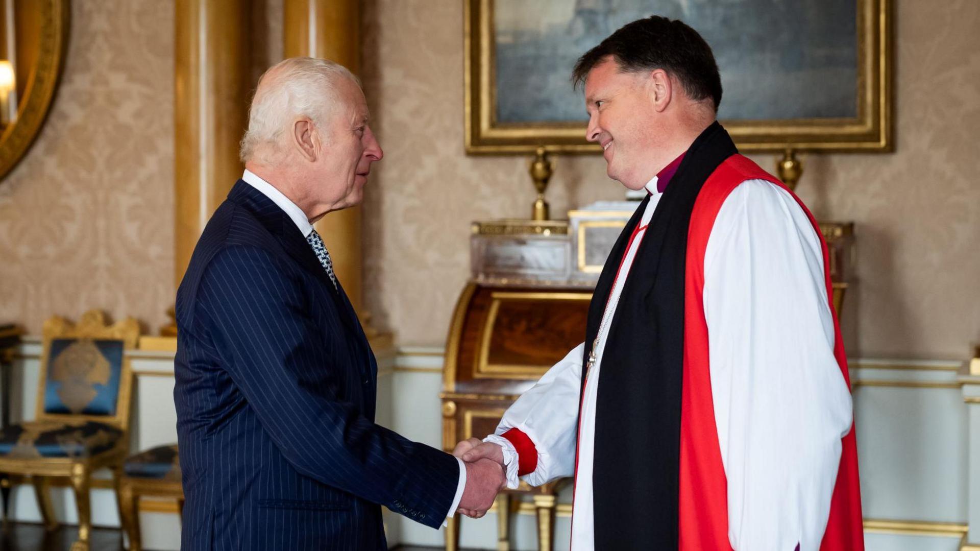 King Charles shaking the hand of the Bishop of Norwich, the Right Reverend Graham Usher. They are inside the 1844 room at Buckingham Palace, which has a piano and chairs in front of a painting on the wall. The pair are looking at each other with friendly expressions as they shake hands. Charles, who has short grey hair, is wearing a navy suit. Bishop Usher is wearing his church attire, consisting of white, black and red robes.