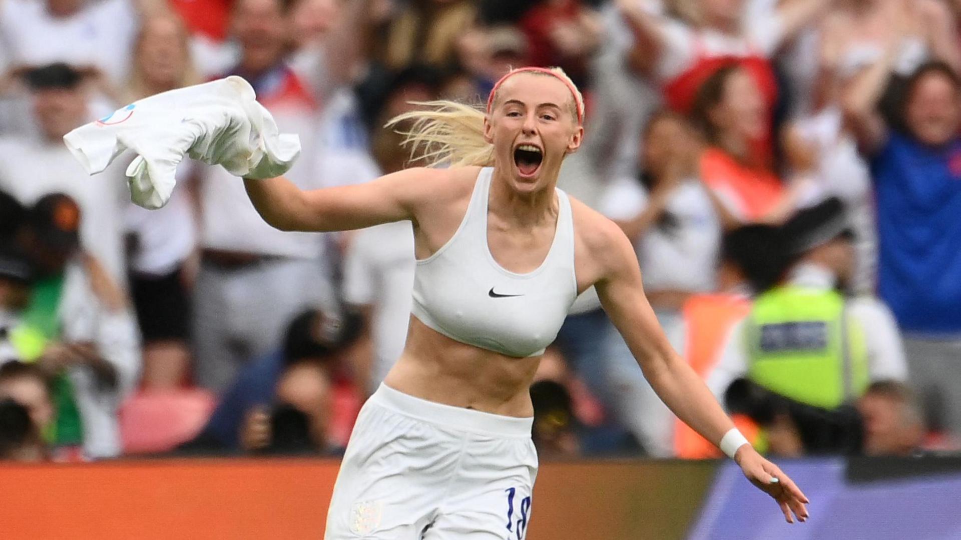 A female footballer celebrates after scoring a goal. The player, who has blonde hair, is holding her white shirt above her head. She is wearing a white sports bra and white shorts. A crowd wearing red, white and blue clothing are visible in the background.