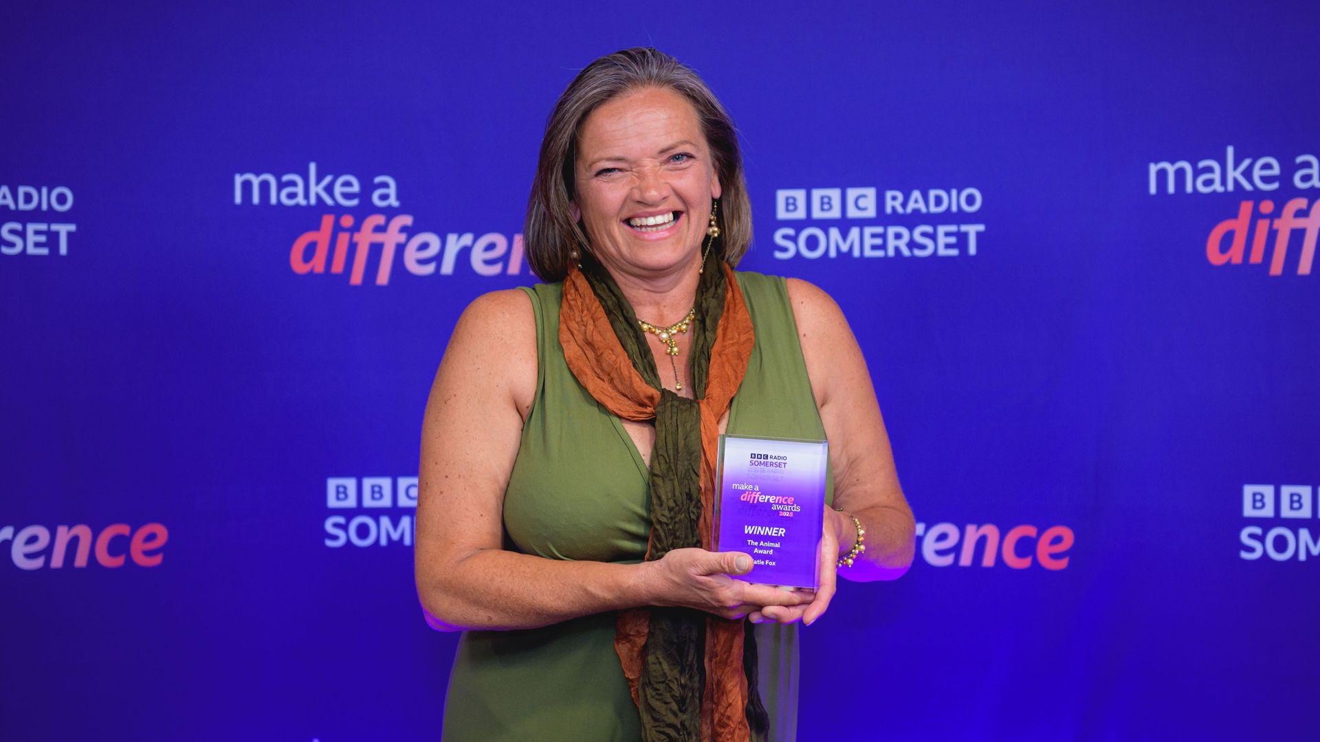 A woman wearing green vest and orange scarf smiles at camera while holding trophy in front of her. Behind her is the promotion wall with "make a difference" and "BBC Somerset" written in pale text on a blue background in multiple places