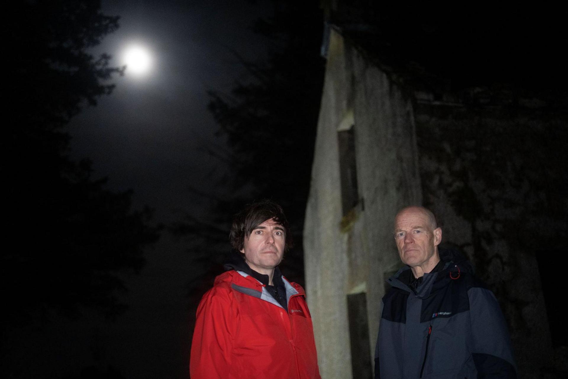 Danny and Phil are standing in the dark outside the ruin of Luibeilt Lodge. A full moon glows behind cloud in the night sky.