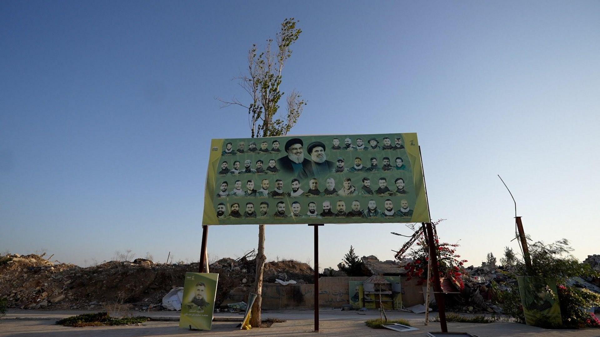 A large green billboard on stilts displays images of men on a green background. Behind the billboard is rubble and destroyed buildings.