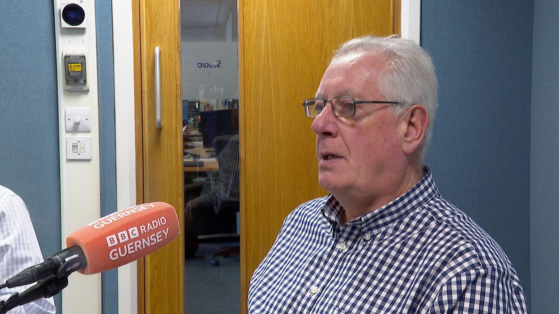 A man in a blue and white checked shirt and an orange microphone with the words BBC Radio Guernsey on it. The man, Paul Luxon, has grey hair and is wearing black rectangular rimmed glasses. 