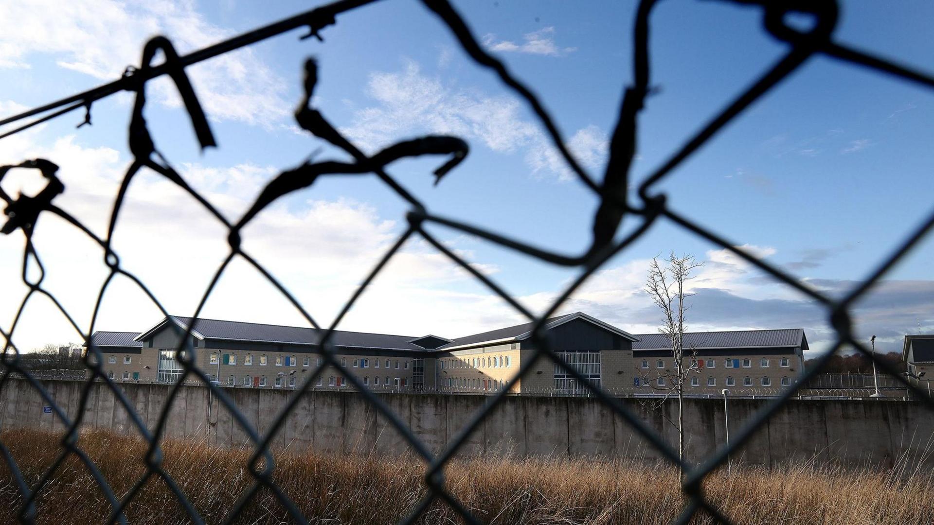 A chain link fence in the foreground with a prison wall and prison building in the background at Low Moss 