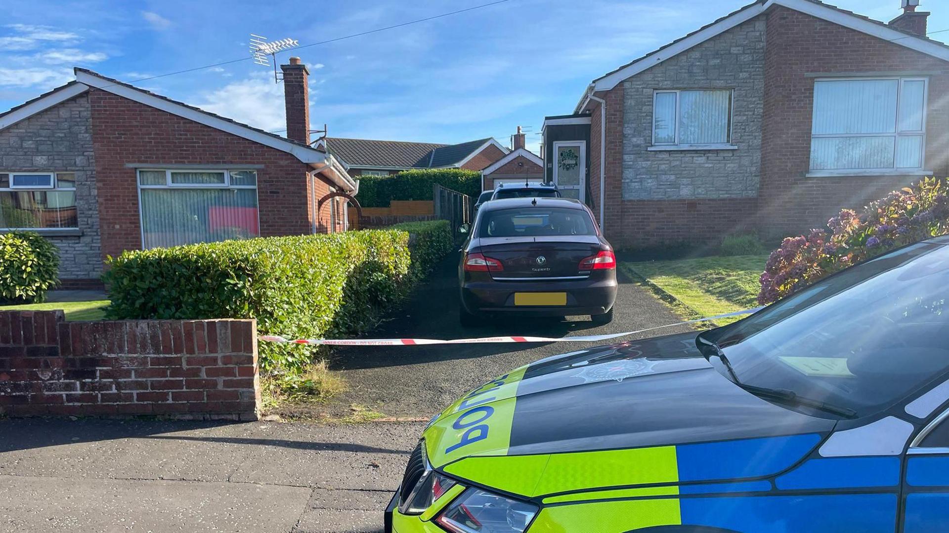 Detached bungalow with two cars in the driveway and police tape across the drive. A police car sits outside the house.