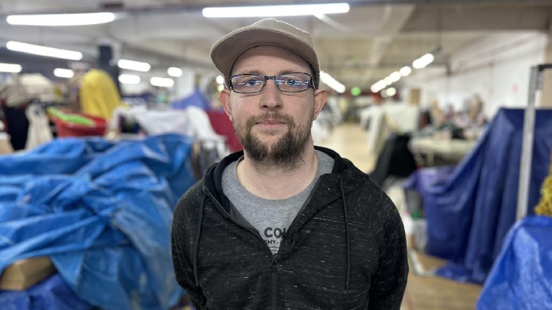 Alec Whitworth, in a hooded top and wearing a baseball camera, looking at the camera while standing in strip lit hall filled with assorted goods