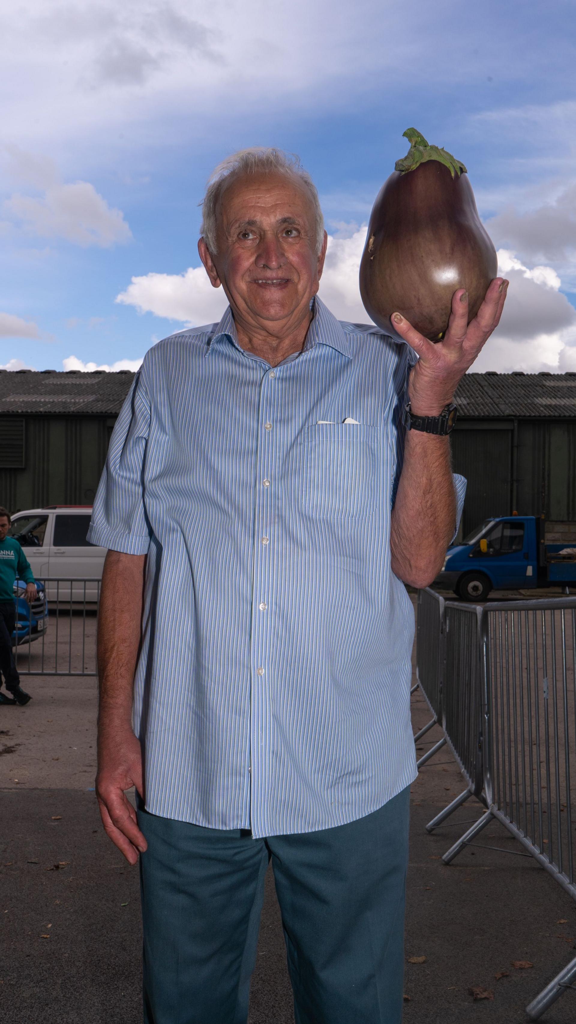 A man holds an enormous aubergine, bright blue clouded sky behind him. 