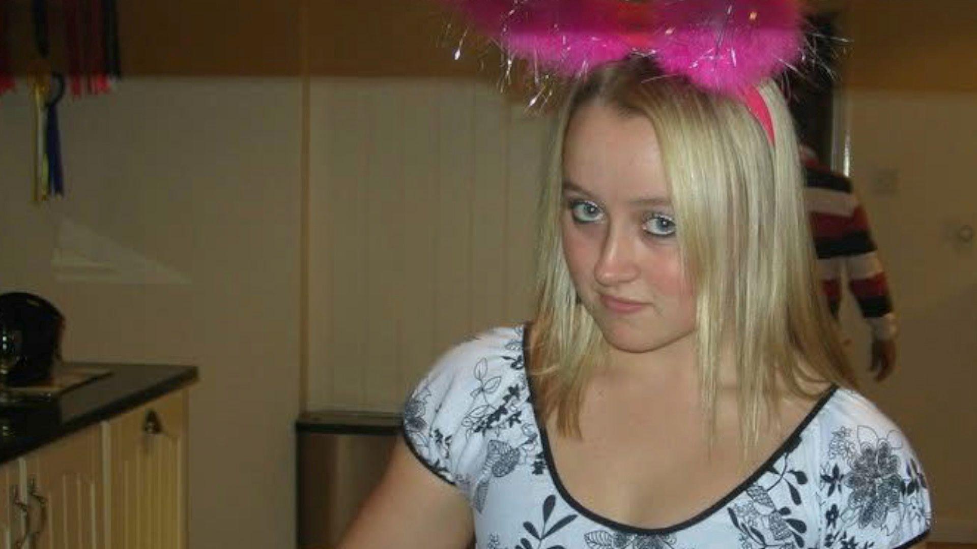 Ella is looking at the camera, she has shoulder length blonde hair and wears a bright pink feather party headband with a short sleeved white and grey floral print t shirt.