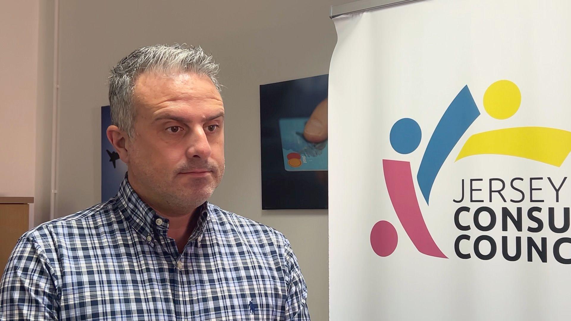 Carl Walker is wearing a blue and white chequered shirt. He is stood next to a Jersey Consumer Council sign. 