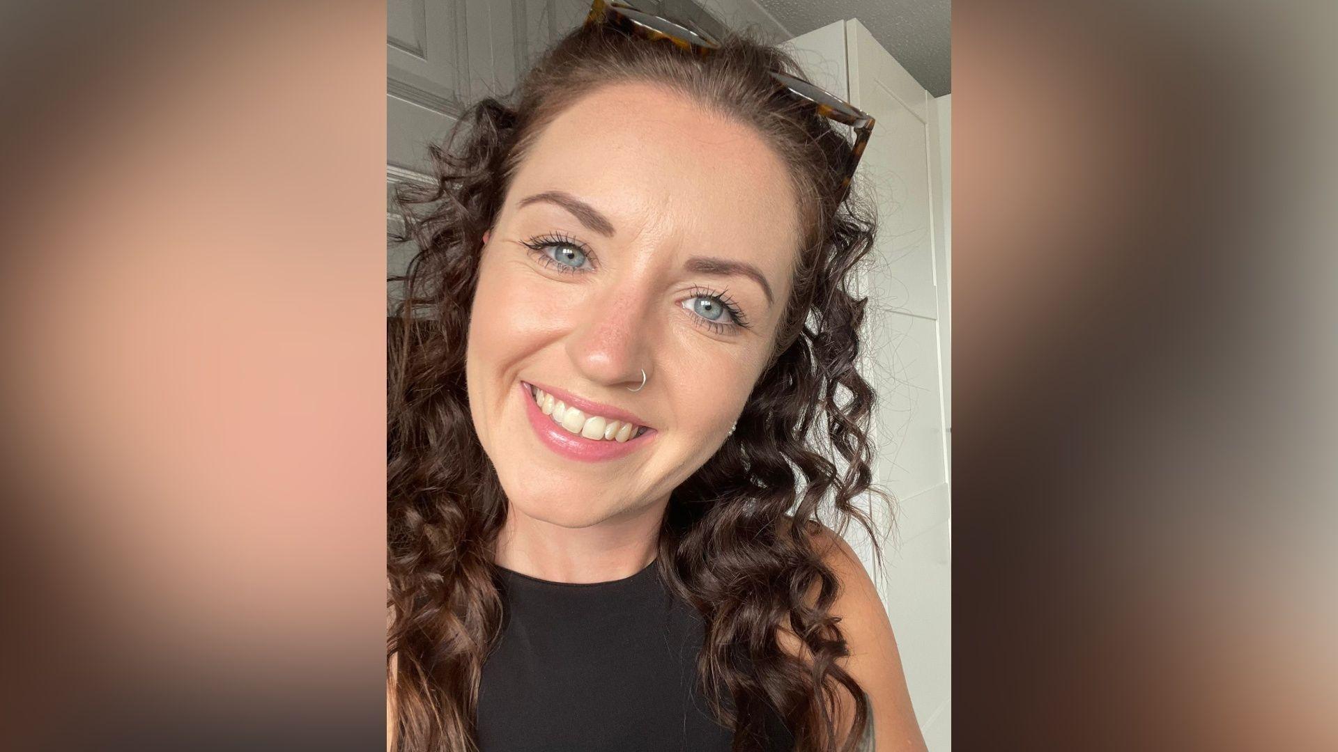 A selfie-style image of Faye Ramsey who is smiling at the camera. She has long brown hair which is curly and also has a nose ring on her right hand nostril. Her glasses rest on the top of her head and she is wearing a black sleeveless top.