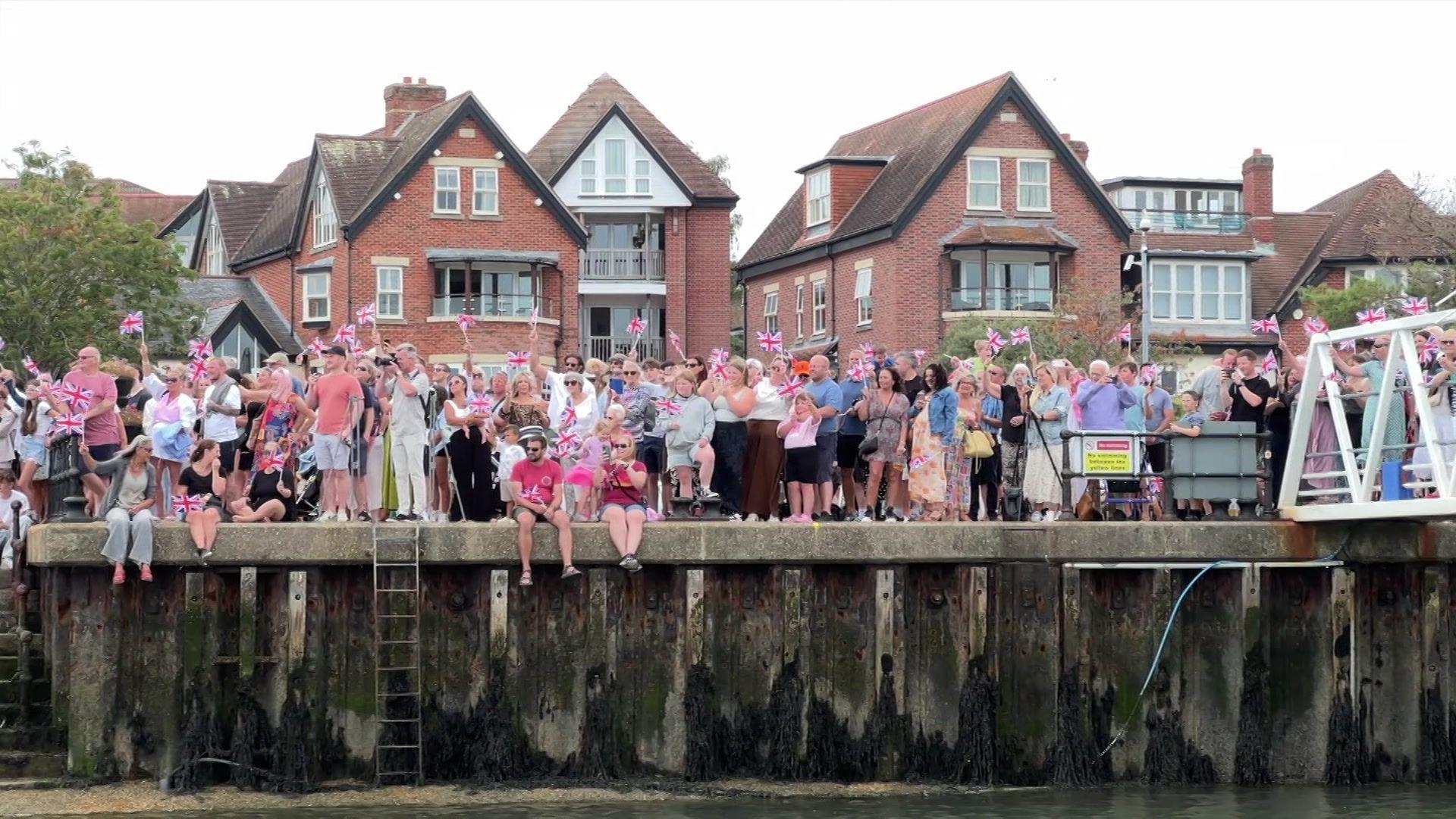 A crowd of supporters awaiting the team at Hamble. Many are waving British flags. Some are dangling their legs over the edge of the wall.