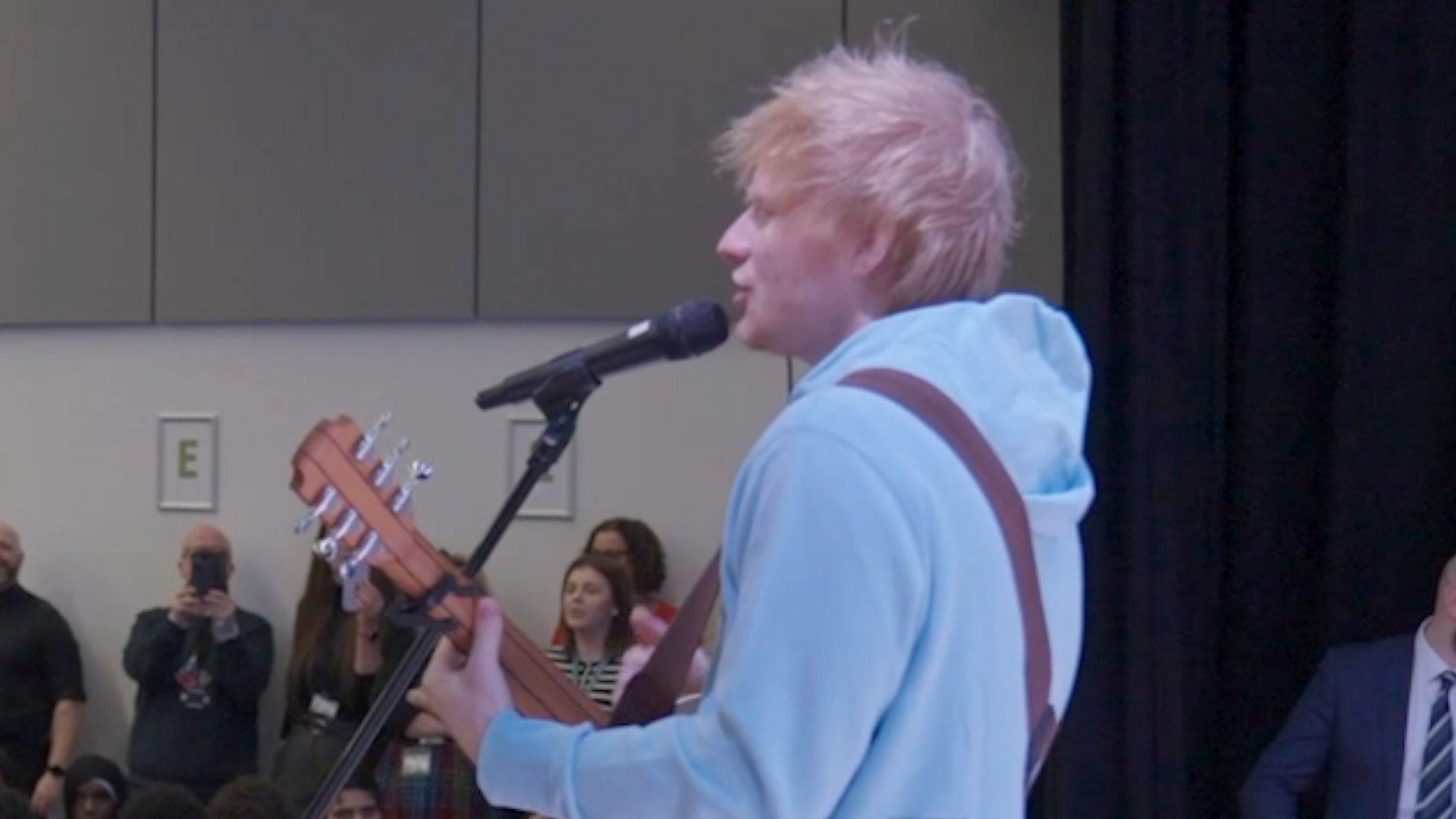 Ed Sheeran in front of a mic and holding a guitar. He is singing