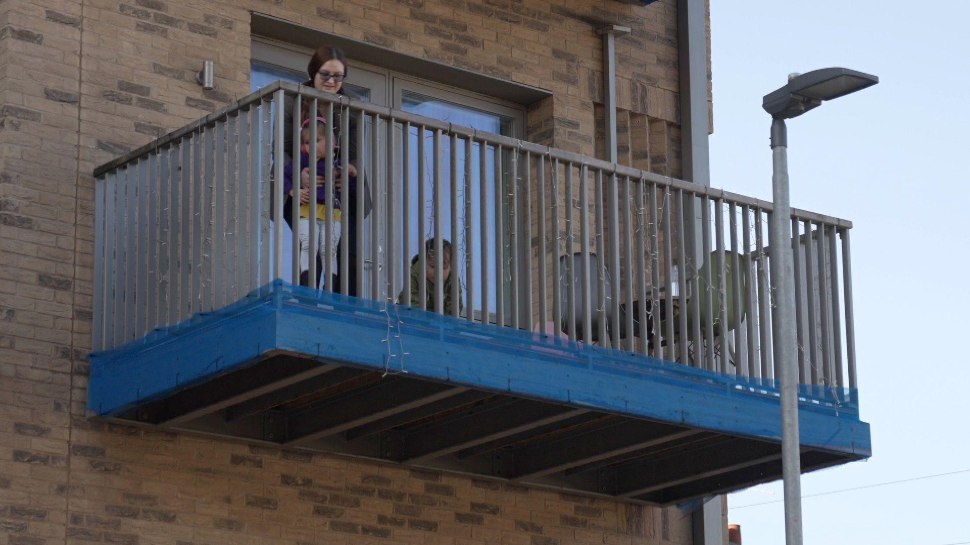 Woman stands alone on a first-floor balcony alongside her children playing with a blue safety covering at the base.